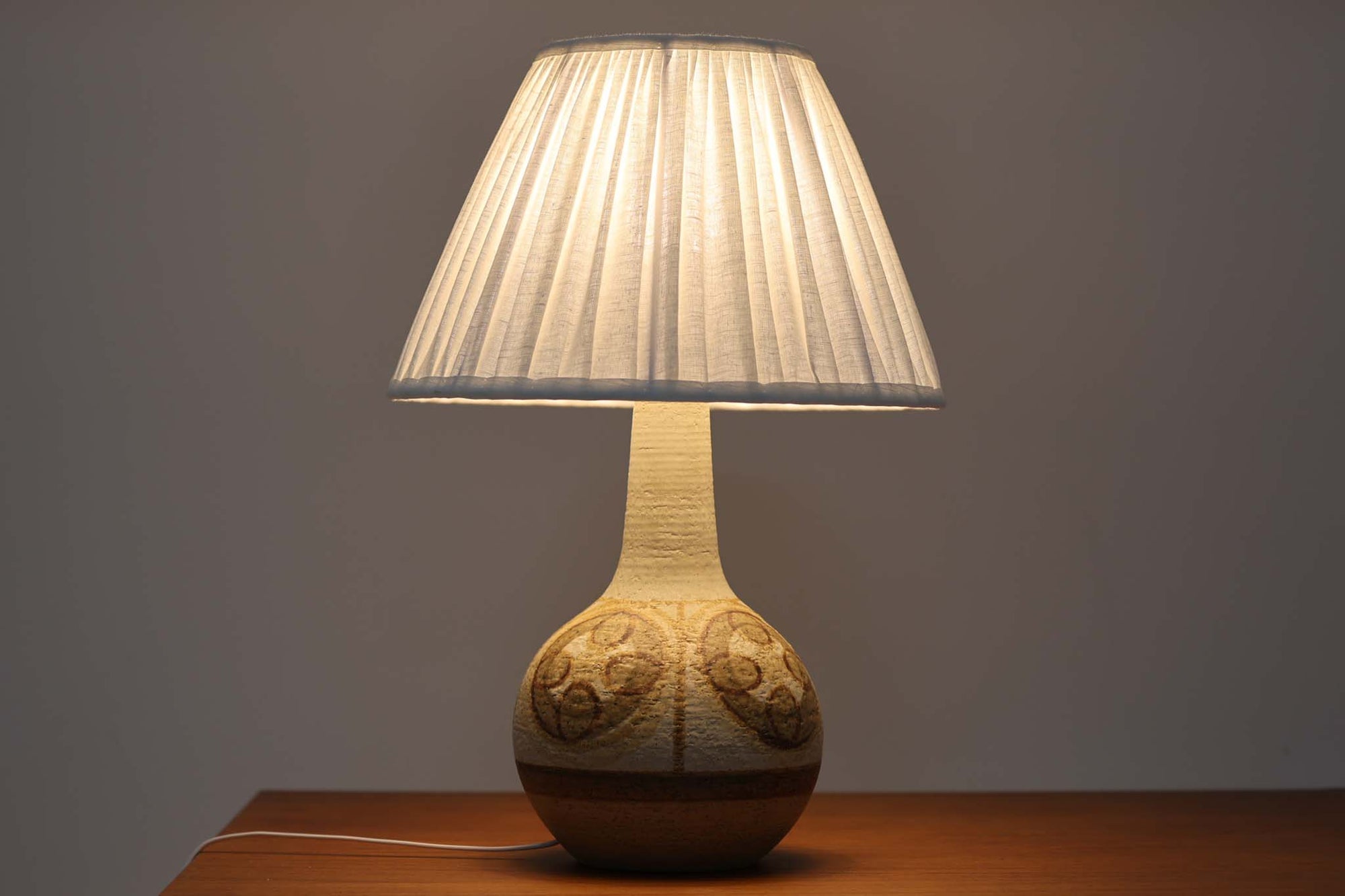 デンマーク・ボーンホルム島の名窯 SOHOLM（スーホルム）によるヴィンテージのテーブルランプです。ふっくらとしたボディに施された幾何学模様が印象的で、素朴であたたかみのある釉薬の風合いが魅力。北欧らしいナチュラルな空気感を空間に添えてくれます。高さは約48cmとやや大きめで、しっかりとした存在感がありながらも落ち着いた佇まいです。シェードは新品のリネン製に交換済み。やさしい光がファブリック越しにふんわりと広がり、ベッドサイドやリビングのアクセントにぴったりです。北欧陶器ならではの土の質感と落ち着いた色彩をお楽しみください。