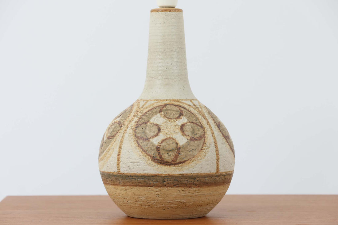 デンマーク・ボーンホルム島の名窯 Soholm（スーホルム）によるヴィンテージのテーブルランプです。ふっくらとしたボディに施された幾何学模様が印象的で、素朴であたたかみのある釉薬の風合いが魅力。北欧らしいナチュラルな空気感を空間に添えてくれます。高さは約48cmとやや大きめで、しっかりとした存在感がありながらも落ち着いた佇まいです。シェードは新品のリネン製に交換済み。やさしい光がファブリック越しにふんわりと広がり、ベッドサイドやリビングのアクセントにぴったりです。北欧陶器ならではの土の質感と落ち着いた色彩をお楽しみください。