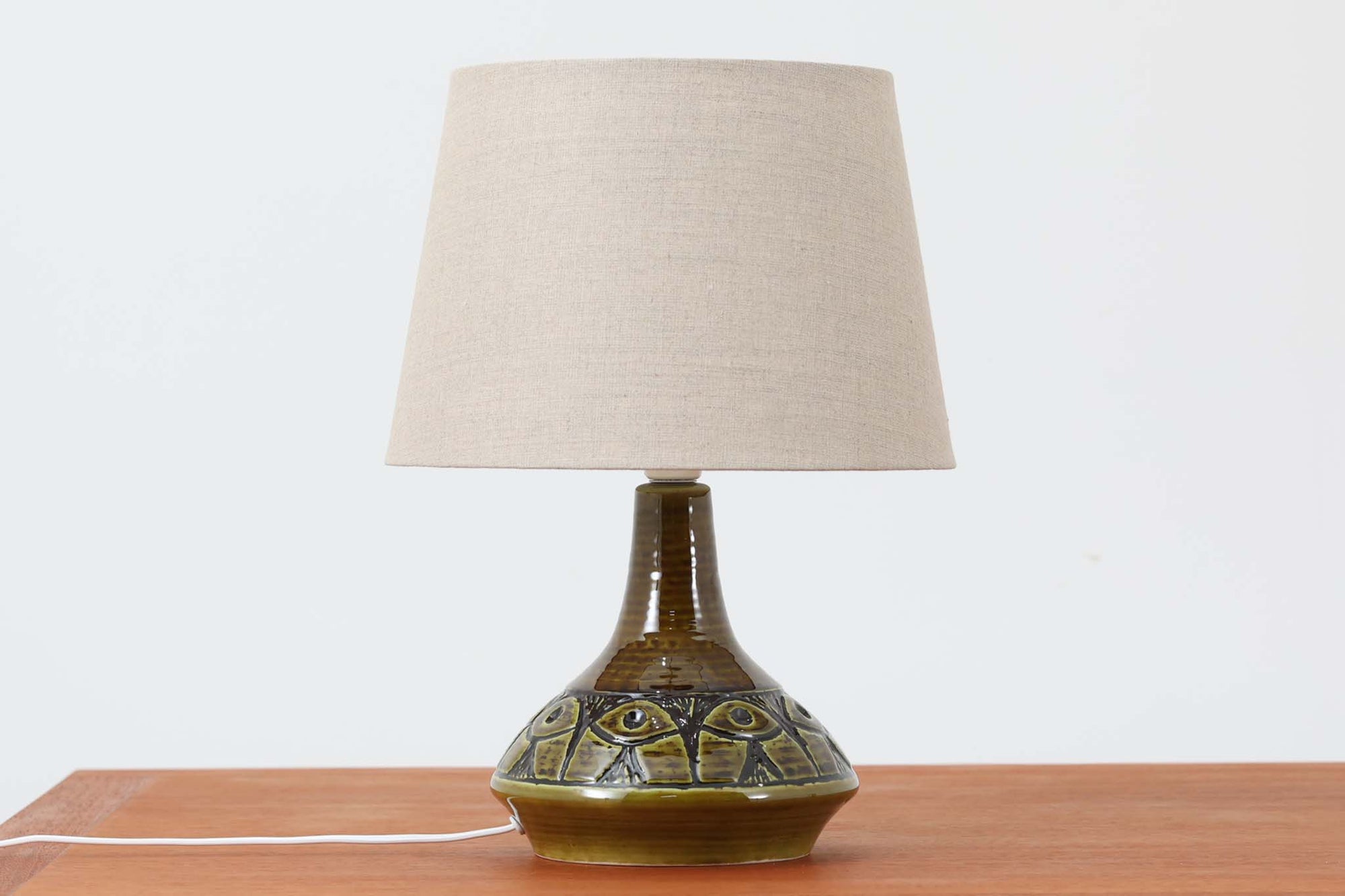 デンマーク・ボーンホルム島の陶器工房SOHOLMによるテーブルランプ。陶肌に浮かび上がる幾何学模様が印象的で、まるで目のようなユニークな装飾が施されています。艶のあるグリーンの釉薬が奥行きを感じさせ、どこか神秘的な雰囲気を漂わせます。シェードは新品のリネン製で、ベースとの相性も良く、空間に柔らかな光を添えてくれます。底面には「SOHOLM DENmARK」のサインが確認できます。北欧ヴィンテージならではの存在感ある一点物です。