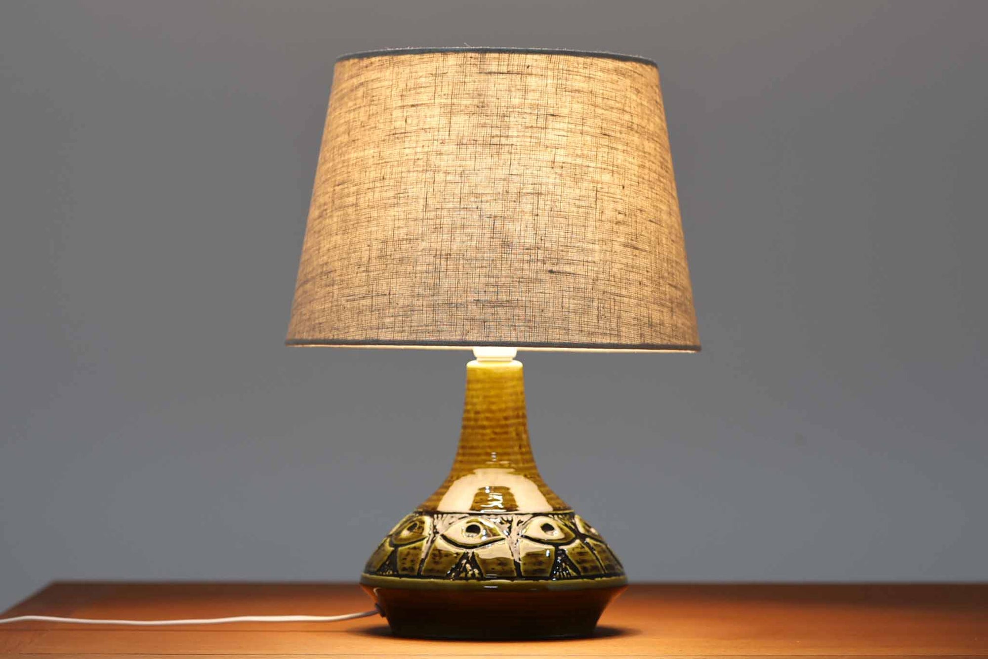 デンマーク・ボーンホルム島の陶器工房SOHOLMによるテーブルランプ。陶肌に浮かび上がる幾何学模様が印象的で、まるで目のようなユニークな装飾が施されています。艶のあるグリーンの釉薬が奥行きを感じさせ、どこか神秘的な雰囲気を漂わせます。シェードは新品のリネン製で、ベースとの相性も良く、空間に柔らかな光を添えてくれます。底面には「SOHOLM DENmARK」のサインが確認できます。北欧ヴィンテージならではの存在感ある一点物です。