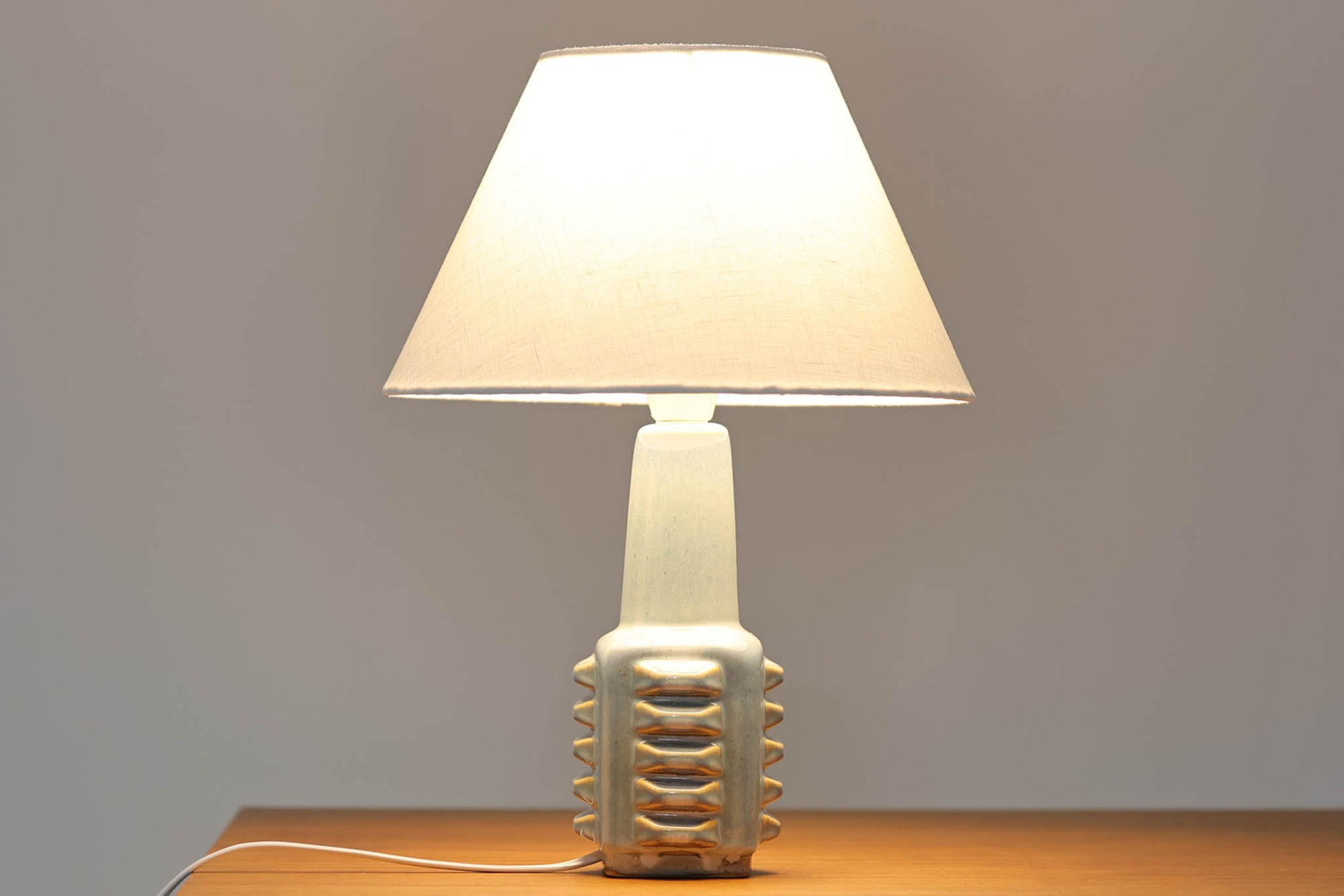 デンマーク・ボーンホルム島の陶器メーカーSOHOLM（スーホルム）で製作されたテーブルランプ。デザイナーはEinar Johansen（アイナー・ヨハンセン）。立体的なレリーフ模様と、ブルーグレーに淡くアイスブルーが重なる美しい釉薬の表情が特徴です。ユニークなフォルムも印象的で、置くだけで空間に存在感をもたらします。シェードは新品のリネン製を合わせています。北欧ヴィンテージらしい温もりと洗練を兼ね備えた一点です。