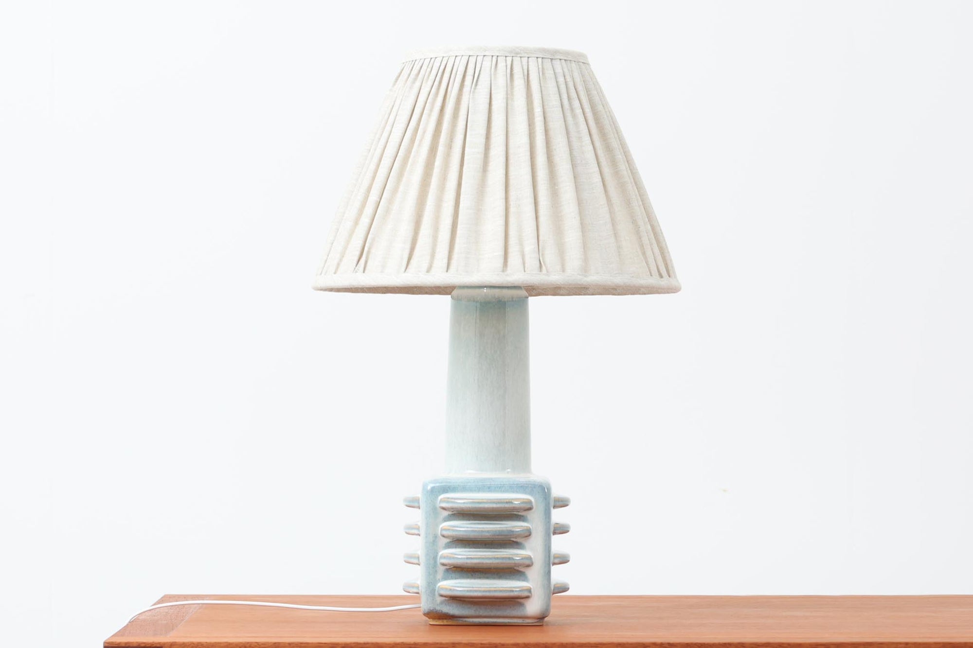 デンマーク・ボーンホルム島の陶器メーカー SOHOLM（スーホルム）で製作されたテーブルランプ。デザイナーは Einar Johansen（アイナー・ヨハンセン）。立体的なレリーフ模様と、ブルーグレーに淡くアイスブルーが重なる美しい釉薬の表情が特徴です。ユニークなフォルムも印象的で、置くだけで空間に存在感をもたらします。高さは約51cmとやや大きめで、サイドボードやコンソールの上などに置くと程よいボリューム感が生まれます。シェードは新品のリネン製を合わせています。北欧ヴィンテージらしい温もりと洗練を兼ね備えた一点です。