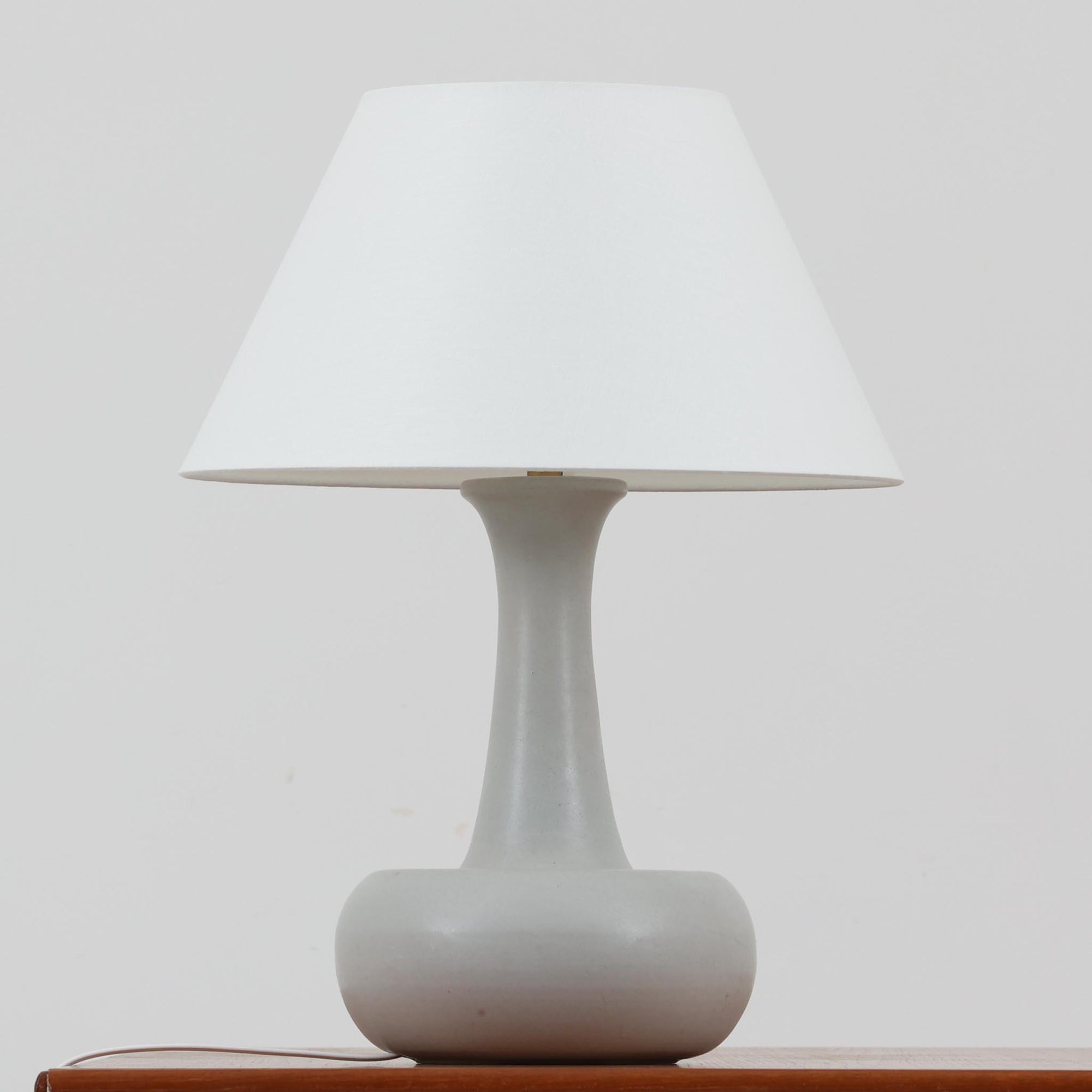 デンマークの照明ブランド Le Klint（レ・クリント）によるテーブルランプで、デザイナーは Ole Boggild（オーレ・ボギル）です。シェードは Le Klint 社製ではありませんが、新品のリネン製プリーツシェードを合わせており、全体として上品で柔らかな印象に仕上がっています。丸みを帯びた陶磁器ベースに、すらりと伸びるネックの造形が美しく、北欧デザインらしい穏やかな佇まいを感じさせます。ややグレーがかったマットな質感が上品で、リネンシェードを通した柔らかな光が空間を優しく包み込みます。シンプルながら存在感があり、リビングやベッドサイドなどさまざまなシーンでお使いいただけます。北欧の上質な灯りをお楽しみいただけるビンテージランプです。