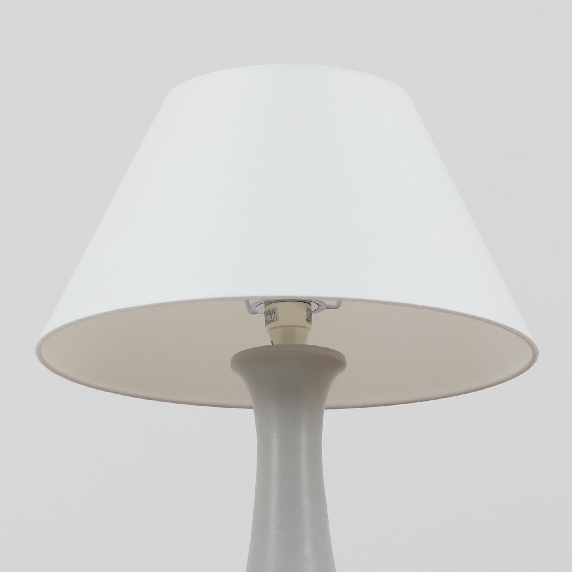 デンマークの照明ブランド Le Klint（レ・クリント）によるテーブルランプで、デザイナーは Ole Boggild（オーレ・ボギル）です。シェードは Le Klint 社製ではありませんが、新品のリネン製プリーツシェードを合わせており、全体として上品で柔らかな印象に仕上がっています。丸みを帯びた陶磁器ベースに、すらりと伸びるネックの造形が美しく、北欧デザインらしい穏やかな佇まいを感じさせます。ややグレーがかったマットな質感が上品で、リネンシェードを通した柔らかな光が空間を優しく包み込みます。シンプルながら存在感があり、リビングやベッドサイドなどさまざまなシーンでお使いいただけます。北欧の上質な灯りをお楽しみいただけるビンテージランプです。