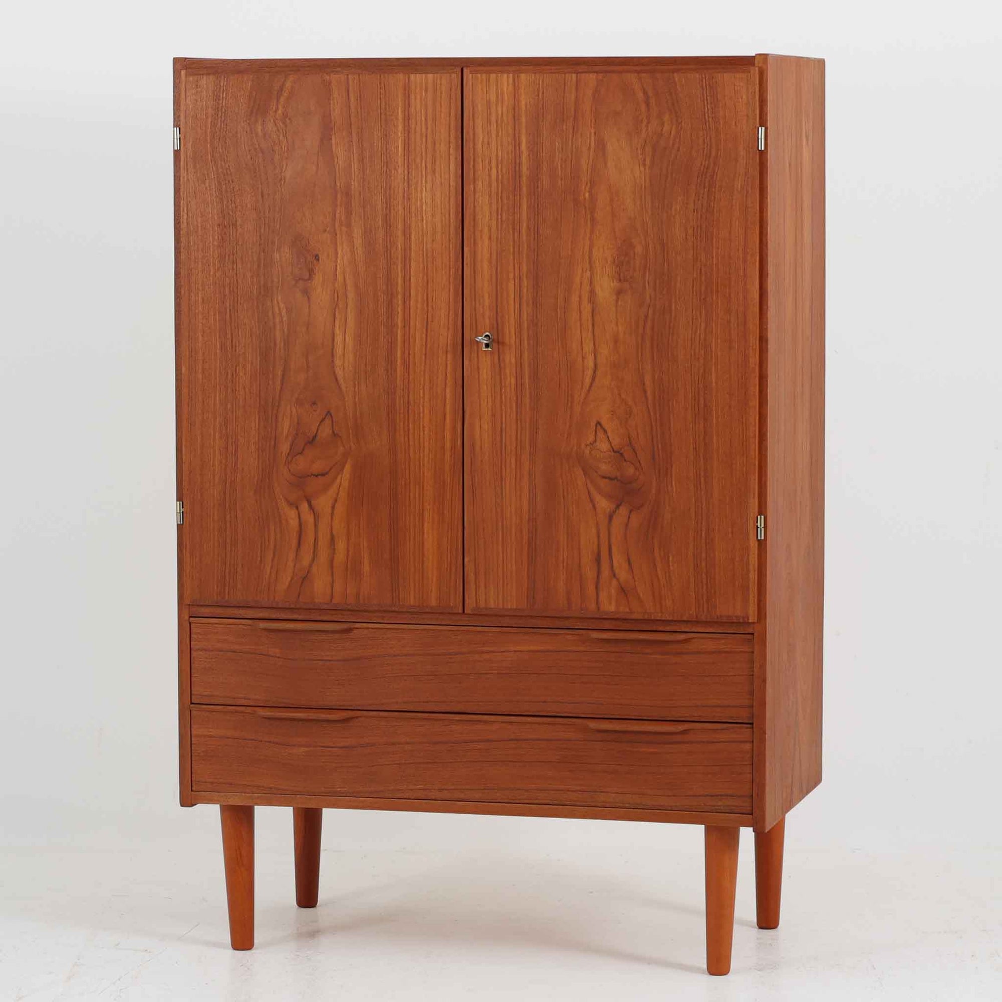 デンマーク製の収納キャビネットです。上段には観音開きの扉内に可動棚が設けられており、食器や書類をすっきりと整理できます。内部まで丁寧に仕上げられており、扉を開けてお気に入りの器やオブジェを飾る“見せる収納”としても楽しめます。扉には鍵が付属しており、大切なものを安全に保管することも可能です。下段には2杯の引き出しが備わり、小物類や日用品の収納にも便利。チーク材の豊かな木目と深みのある色合いが美しく、北欧家具らしい温もりを感じさせます。脚部のテーパー形状が全体を軽やかに見せ、シンプルながら上質な存在感を放つ一台です。