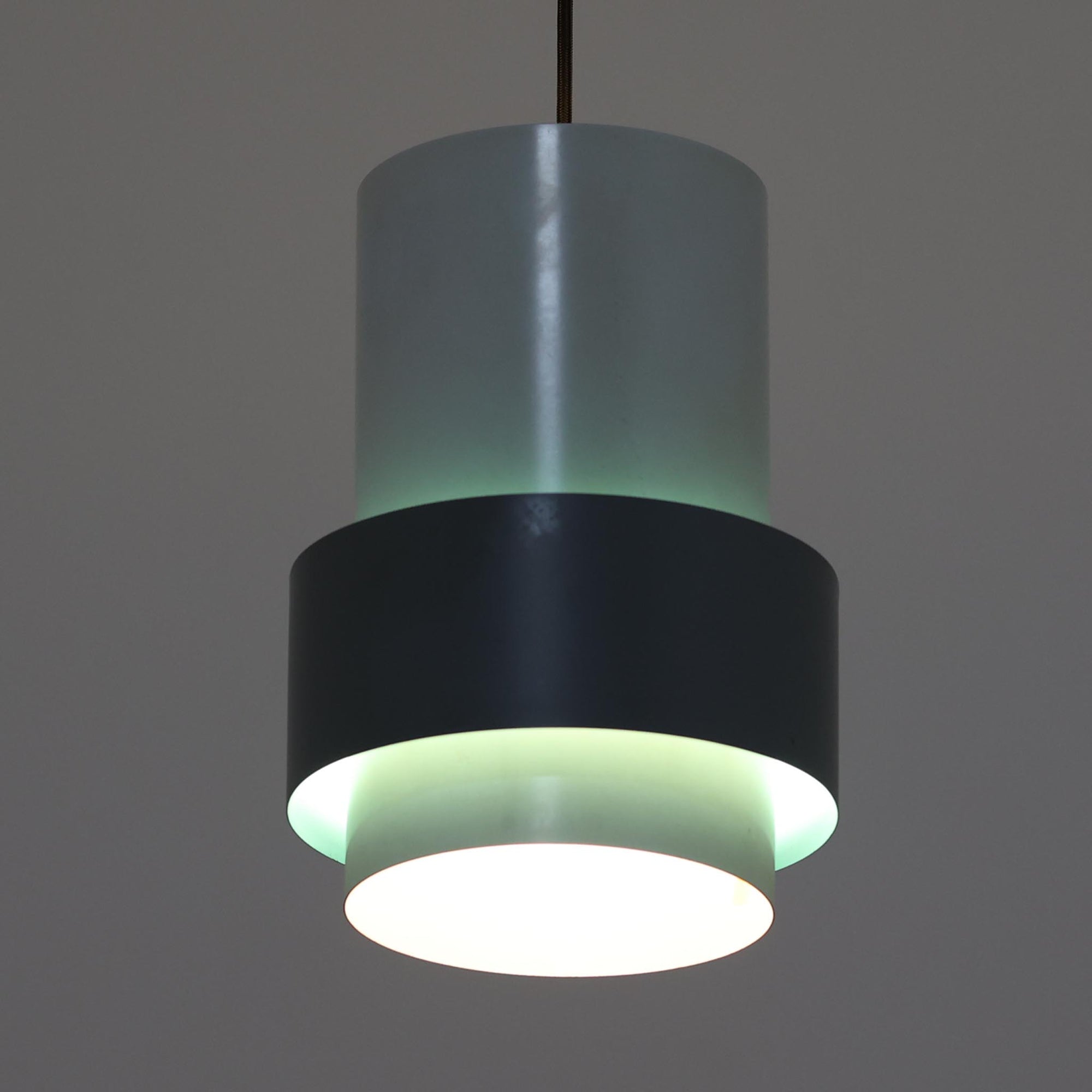Jo HammerborgがFog &amp; Morup社のためにデザインした「Central」ペンダントライトです。直径23cmのコンパクトなサイズながら、スチール製の二重シリンダー構造が特徴的で、点灯時には上下から光が広がり独特の陰影を演出します。外側のグレーと内側のホワイトがコントラストを生み、点灯時にはほのかなグリーンの光が漏れ出す美しいデザインです。ダイニングや小スペースに適したサイズで、北欧モダンのエッセンスを感じさせるビンテージ照明です。シェードの内側に施されたグリーンの塗装が、点灯時に柔らかなニュアンスを加え、空間に独特の雰囲気を演出します。