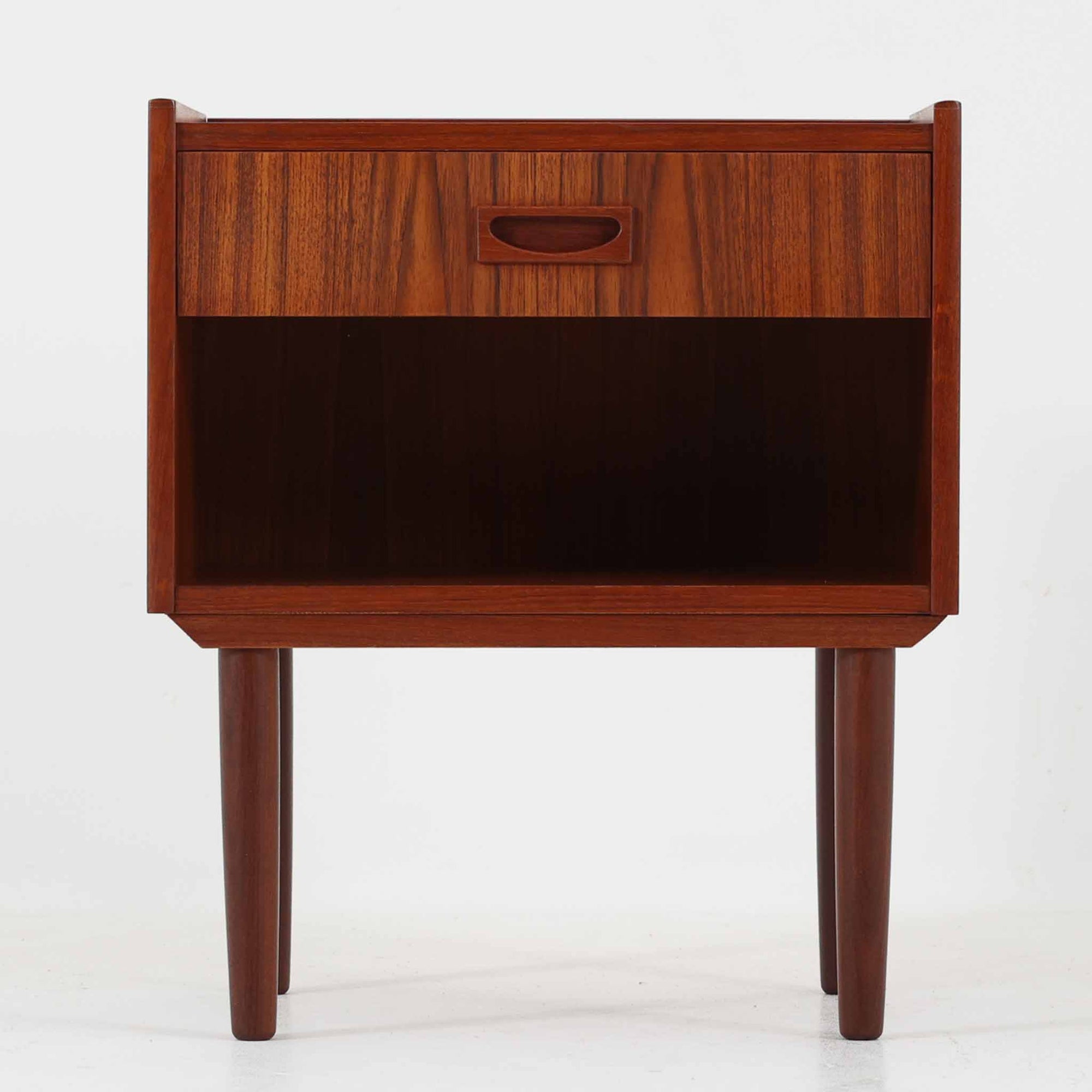 チーク材の豊かな木目が美しいこちらのサイドチェストは、北欧ビンテージらしい端正なデザインと実用性を兼ね備えた一台です。天板の縁には立ち上がりが施され、小物の転落を防ぐ工夫がされています。引き出し下のオープンスペースは本や雑貨の収納に便利で、ソファやベッドサイドに置けばサイドテーブルとしても活躍します。丸みを帯びた持ち手やテーパードレッグが柔らかな印象を与え、経年変化による木肌の深みが空間に温もりを添える逸品です。