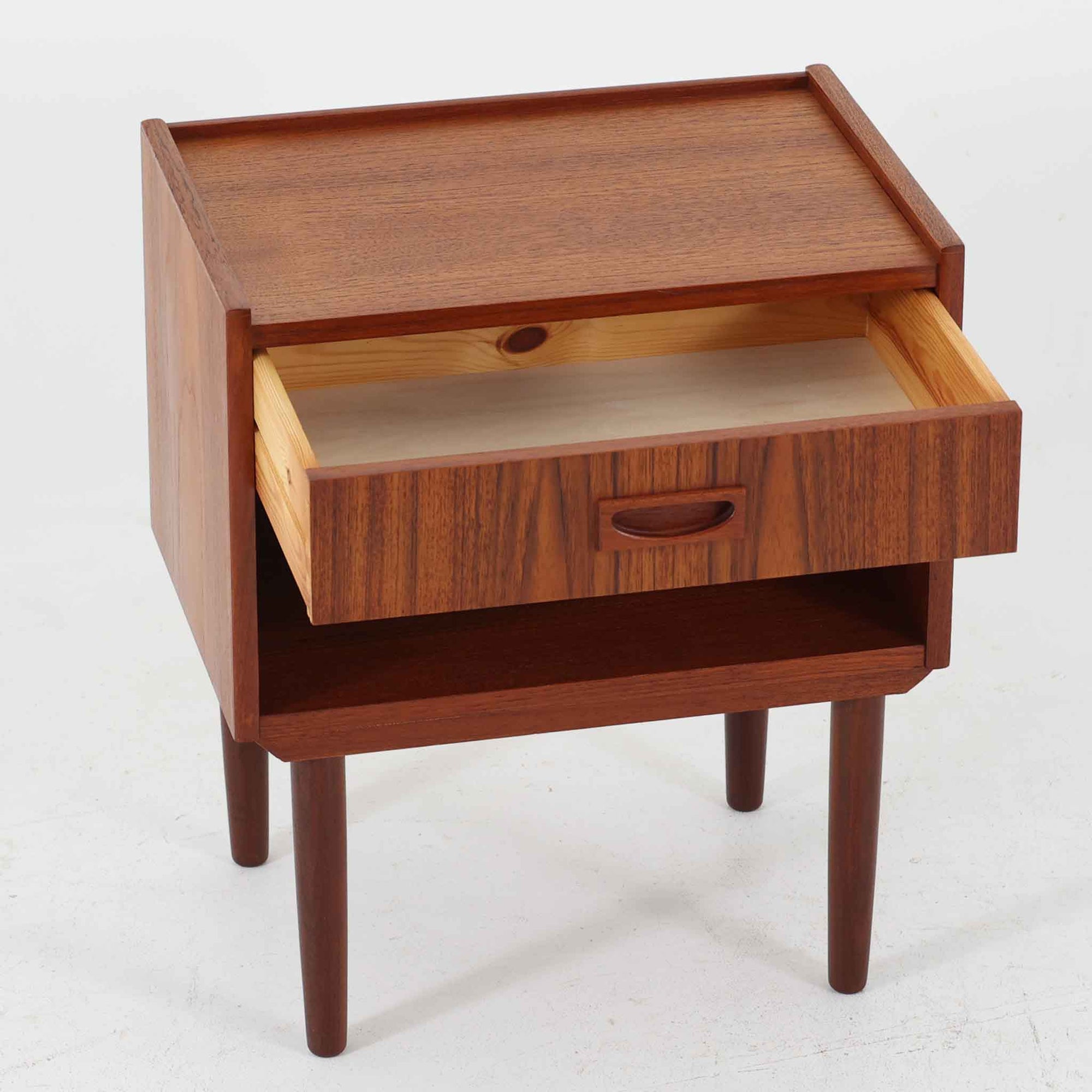 チーク材の豊かな木目が美しいこちらのサイドチェストは、北欧ビンテージらしい端正なデザインと実用性を兼ね備えた一台です。天板の縁には立ち上がりが施され、小物の転落を防ぐ工夫がされています。引き出し下のオープンスペースは本や雑貨の収納に便利で、ソファやベッドサイドに置けばサイドテーブルとしても活躍します。丸みを帯びた持ち手やテーパードレッグが柔らかな印象を与え、経年変化による木肌の深みが空間に温もりを添える逸品です。