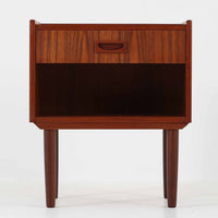 チーク材の豊かな木目が美しいこちらのサイドチェストは、北欧ビンテージらしい端正なデザインと実用性を兼ね備えた一台です。天板の縁には立ち上がりが施され、小物の転落を防ぐ工夫がされています。引き出し下のオープンスペースは本や雑貨の収納に便利で、ソファやベッドサイドに置けばサイドテーブルとしても活躍します。丸みを帯びた持ち手やテーパードレッグが柔らかな印象を与え、経年変化による木肌の深みが空間に温もりを添える逸品です。