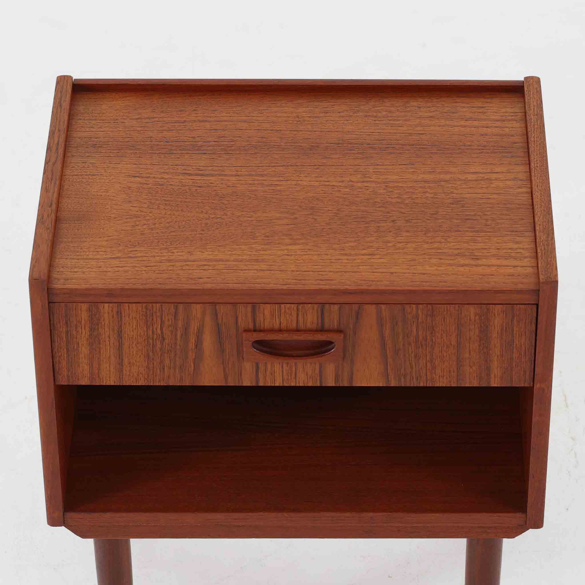 チーク材の豊かな木目が美しいこちらのサイドチェストは、北欧ビンテージらしい端正なデザインと実用性を兼ね備えた一台です。天板の縁には立ち上がりが施され、小物の転落を防ぐ工夫がされています。引き出し下のオープンスペースは本や雑貨の収納に便利で、ソファやベッドサイドに置けばサイドテーブルとしても活躍します。丸みを帯びた持ち手やテーパードレッグが柔らかな印象を与え、経年変化による木肌の深みが空間に温もりを添える逸品です。