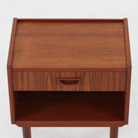 チーク材の豊かな木目が美しいこちらのサイドチェストは、北欧ビンテージらしい端正なデザインと実用性を兼ね備えた一台です。天板の縁には立ち上がりが施され、小物の転落を防ぐ工夫がされています。引き出し下のオープンスペースは本や雑貨の収納に便利で、ソファやベッドサイドに置けばサイドテーブルとしても活躍します。丸みを帯びた持ち手やテーパードレッグが柔らかな印象を与え、経年変化による木肌の深みが空間に温もりを添える逸品です。