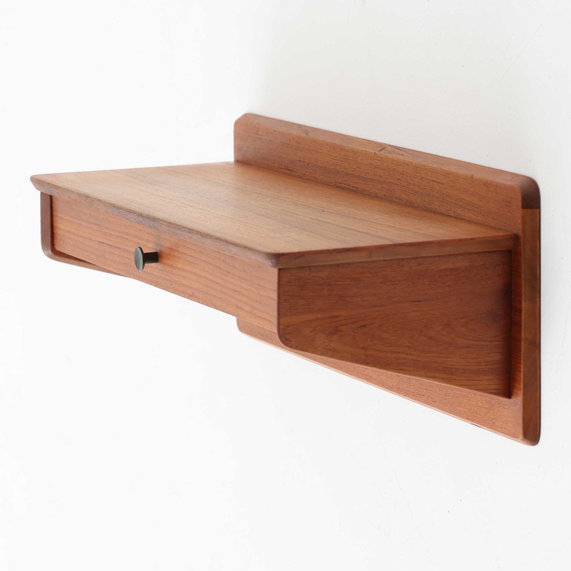 Gunni Omann（グンニ・オーマン）によるデザインで、Omann Jun社から発表されたModel 144ウォールシェルフです。チーク材を使用した上質な仕上がりで、シンプルながらも端正なフォルムと美しい木目が際立ちます。引き出しを備えているため、小物収納にも便利で、ベッドサイドや玄関先など限られたスペースでも活躍します。前板に施された小さなラウンド型の取っ手がデザインのアクセントとなり、北欧家具らしい機能美を感じさせます。経年による風合いが増し、空間に温かみと深みを添えるビンテージの逸品です。