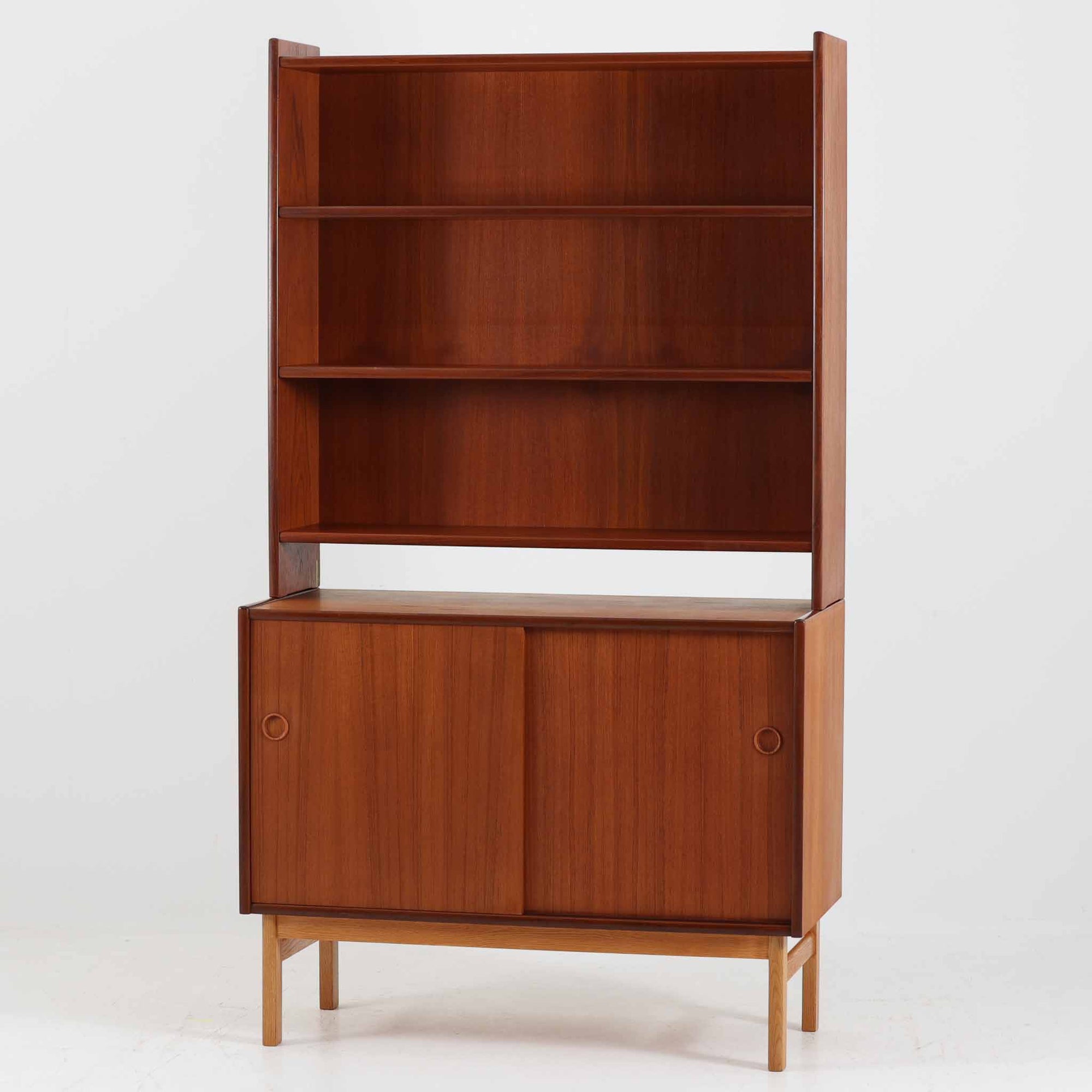 スウェーデン製のディスプレイシェルフで、上段はチーク材の棚板が連なる開放的なディスプレイスペース、下段はオーク材の脚部に支えられたスライド扉収納を備えた北欧ビンテージらしい構成です。丸型の指掛けや直線的なフォルムが端正な印象を与え、見せる収納と隠す収納を兼ね備えた実用性の高い一台です。食器や雑貨、本のディスプレイにも最適で、経年により深みを増した木肌が空間に温かみと上質さを添えます。