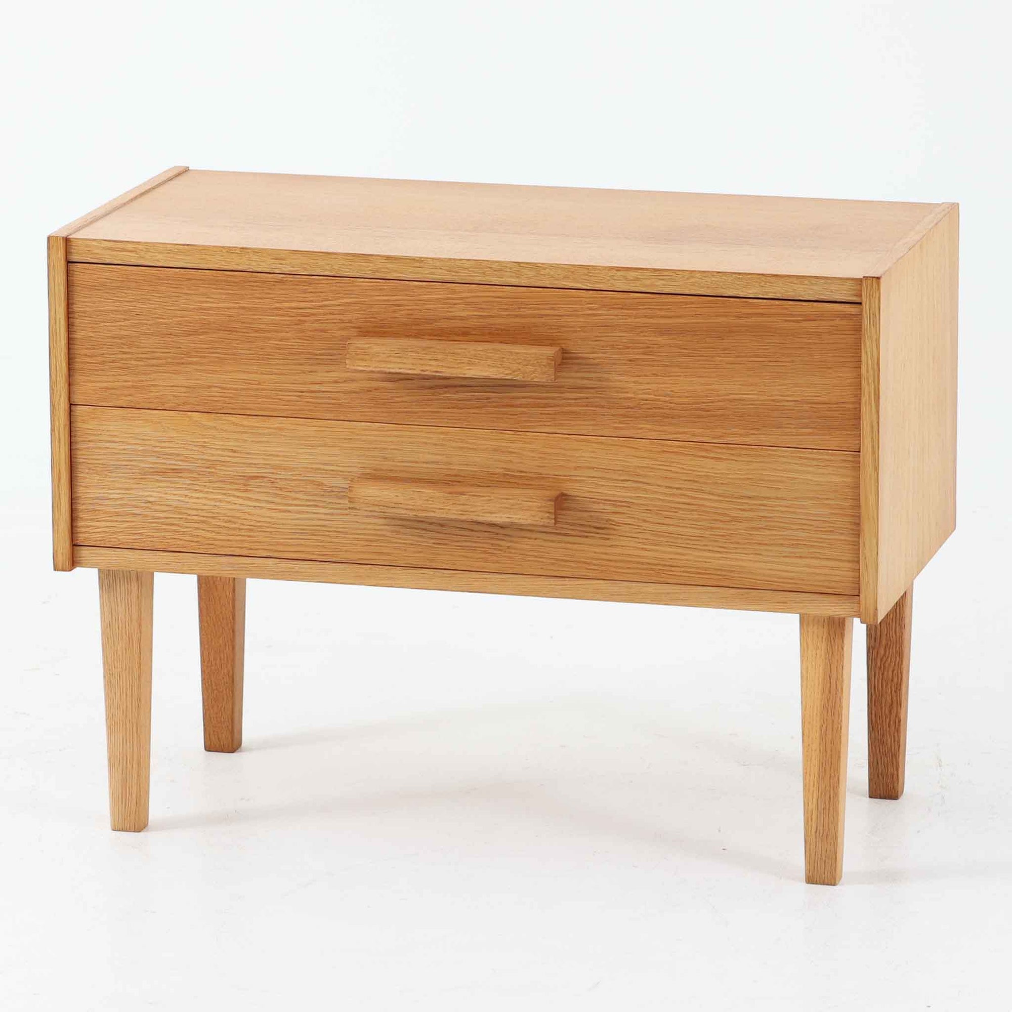 オーク材を用いたこちらの2段チェストは、北欧ビンテージ家具らしいシンプルかつ機能的なデザインが魅力です。直線的なフォルムに対し、細身でテーパーのかかった脚部が軽やかな印象を与え、空間を圧迫せずに置くことができます。幅60cmとコンパクトながら、2杯の引き出しは日用品や小物収納に便利で、ベッドサイドやリビングの補助収納としても最適です。経年により深みを増したオークの木肌が、温もりある雰囲気を演出する一点物です。