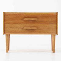 オーク材を用いたこちらの2段チェストは、北欧ビンテージ家具らしいシンプルかつ機能的なデザインが魅力です。直線的なフォルムに対し、細身でテーパーのかかった脚部が軽やかな印象を与え、空間を圧迫せずに置くことができます。幅60cmとコンパクトながら、2杯の引き出しは日用品や小物収納に便利で、ベッドサイドやリビングの補助収納としても最適です。経年により深みを増したオークの木肌が、温もりある雰囲気を演出する一点物です。