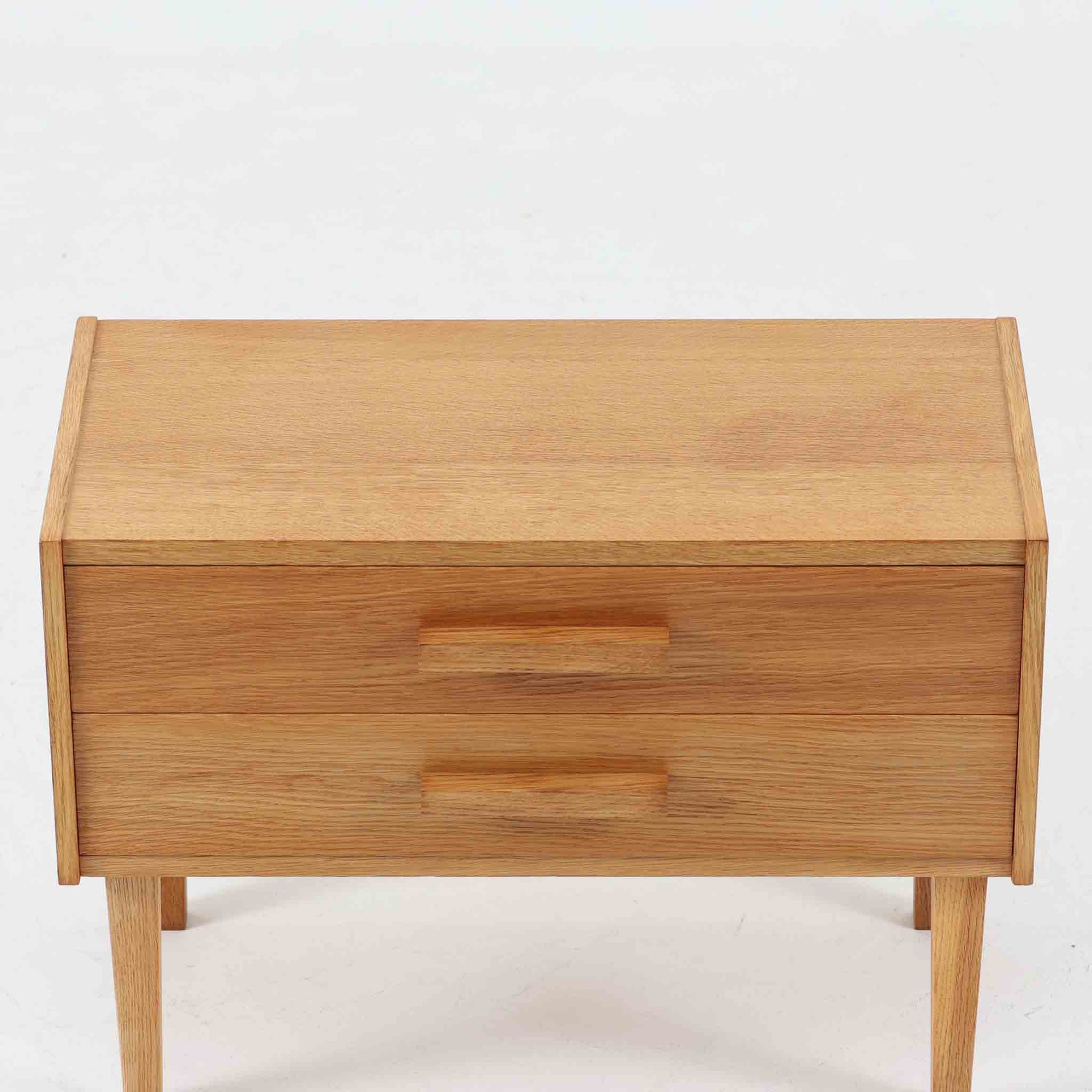 オーク材を用いたこちらの2段チェストは、北欧ビンテージ家具らしいシンプルかつ機能的なデザインが魅力です。直線的なフォルムに対し、細身でテーパーのかかった脚部が軽やかな印象を与え、空間を圧迫せずに置くことができます。幅60cmとコンパクトながら、2杯の引き出しは日用品や小物収納に便利で、ベッドサイドやリビングの補助収納としても最適です。経年により深みを増したオークの木肌が、温もりある雰囲気を演出する一点物です。