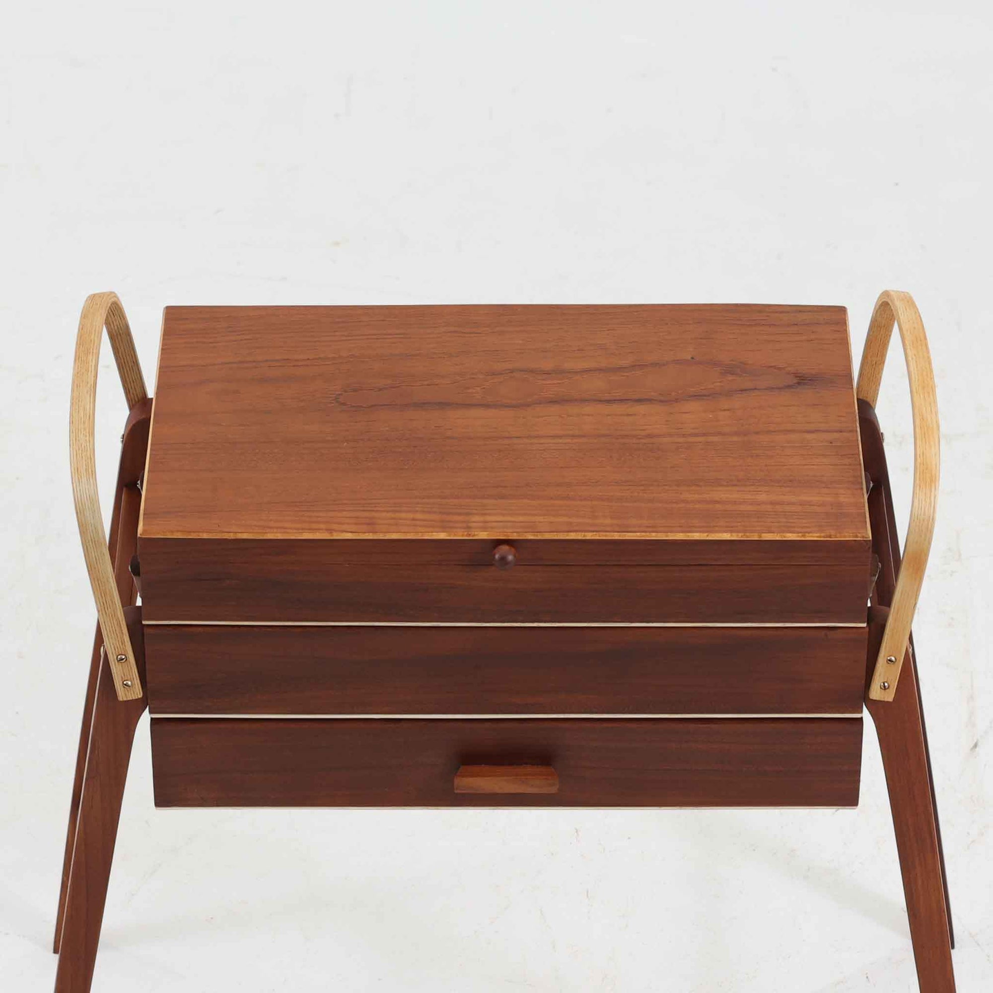 こちらのソーイングボックスは、チーク材とビーチ材を組み合わせた軽やかな構成が魅力の北欧ビンテージ品で、トップのフラップを開くと段違いに展開するトレイと下段の引き出しが現れる優れた収納構造を備え、裁縫道具や小物を効率よく整理できます。柔らかく曲げられた左右のビーチ材の持ち手がアクセントとなり、職人技の感じられる繊細な作りと経年で深まった木肌の風合いが空間に温かな存在感をもたらします。リビングやワークスペースのサイドテーブルとしても実用的にお使いいただけます。