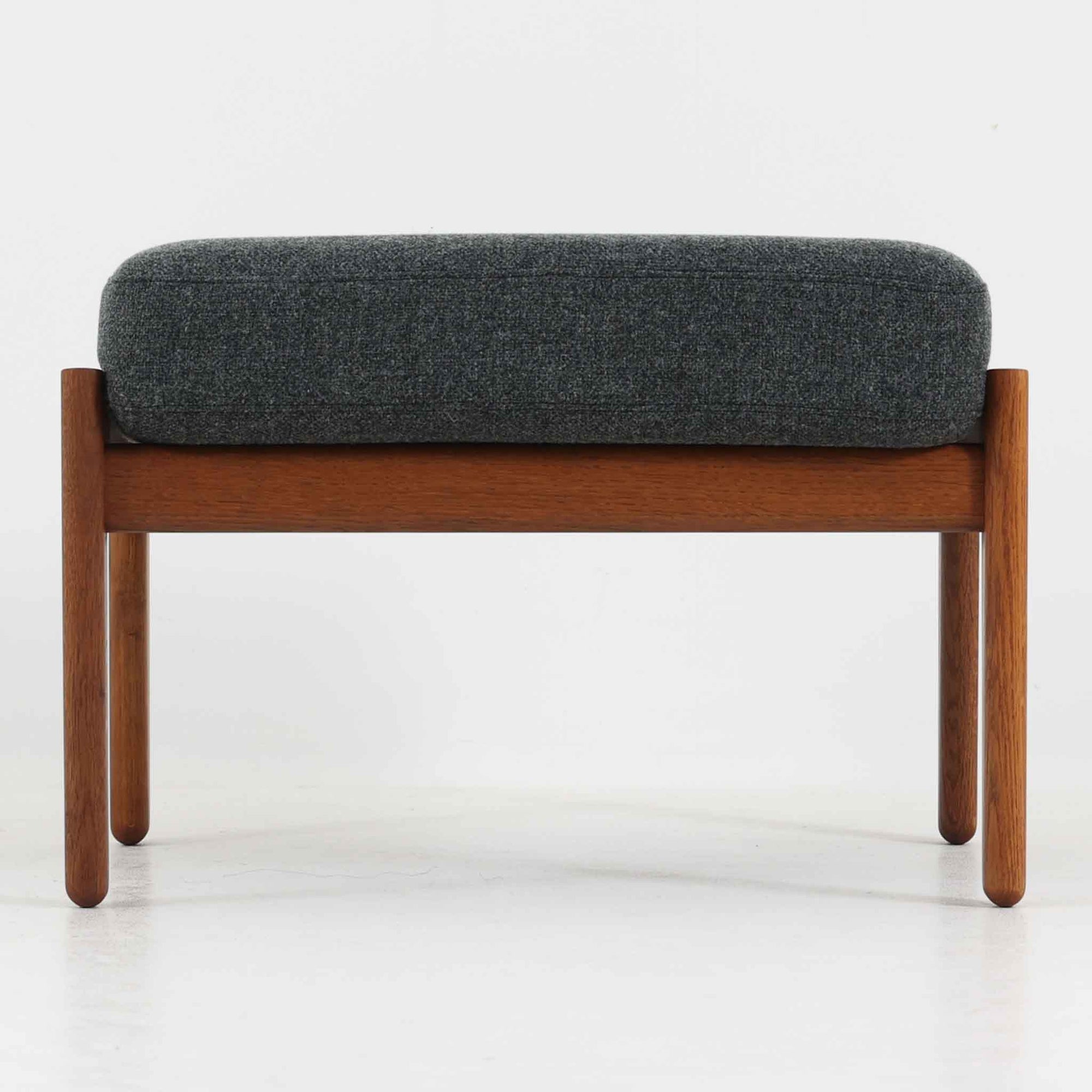 ハンス・J・ウェグナーがデザインし、ゲタマ社から発表されたGE290オットマンは、堅牢なオーク材のフレームと、ウール混紡のざっくりとした風合いを持つダークグレーの張地が美しく調和した北欧ビンテージ家具です。直線的でありながら丸みを帯びた脚部が軽快な印象を与え、ソファと組み合わせてフットレストとして、あるいは単体で腰掛けとしてもお使いいただけます。オーク材の色味は少し濃いめで、本革カバー仕様のビンテージモデルのためクッション内部にスプリングは元々内包されないタイプです。経年によって深まった木肌の風合いと上質な張地が、空間に落ち着いた存在感をもたらす逸品です。