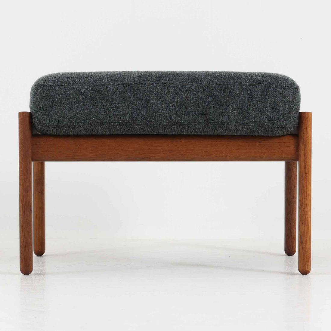 ハンス・J・ウェグナーがデザインし、ゲタマ社から発表されたGE290オットマンは、堅牢なオーク材のフレームと、ウール混紡のざっくりとした風合いを持つダークグレーの張地が美しく調和した北欧ビンテージ家具です。直線的でありながら丸みを帯びた脚部が軽快な印象を与え、ソファと組み合わせてフットレストとして、あるいは単体で腰掛けとしてもお使いいただけます。オーク材の色味は少し濃いめで、本革カバー仕様のビンテージモデルのためクッション内部にスプリングは元々内包されないタイプです。経年によって深まった木肌の風合いと上質な張地が、空間に落ち着いた存在感をもたらす逸品です。