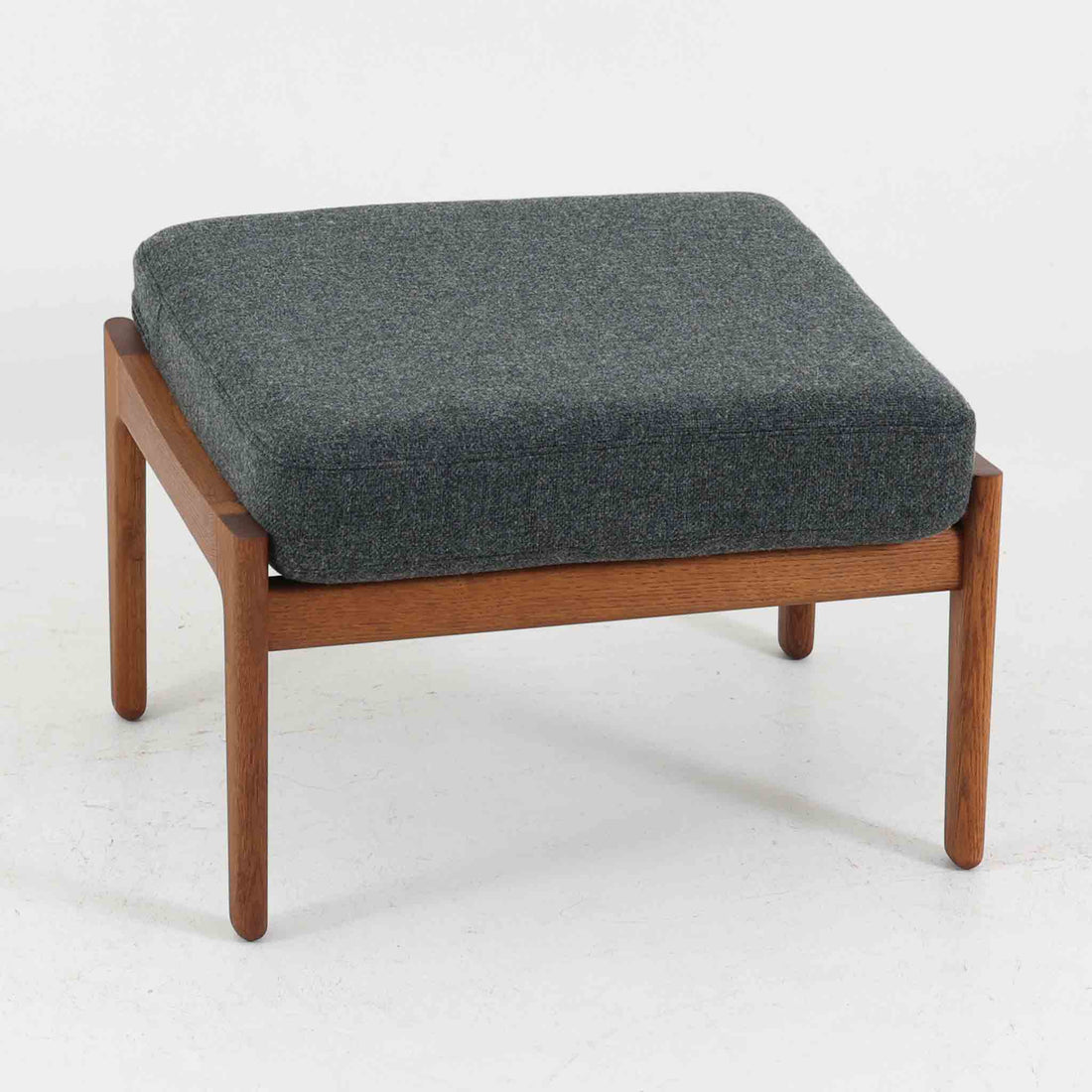 ハンス・J・ウェグナーがデザインし、ゲタマ社から発表されたGE290オットマンは、堅牢なオーク材のフレームと、ウール混紡のざっくりとした風合いを持つダークグレーの張地が美しく調和した北欧ビンテージ家具です。直線的でありながら丸みを帯びた脚部が軽快な印象を与え、ソファと組み合わせてフットレストとして、あるいは単体で腰掛けとしてもお使いいただけます。オーク材の色味は少し濃いめで、本革カバー仕様のビンテージモデルのためクッション内部にスプリングは元々内包されないタイプです。経年によって深まった木肌の風合いと上質な張地が、空間に落ち着いた存在感をもたらす逸品です。