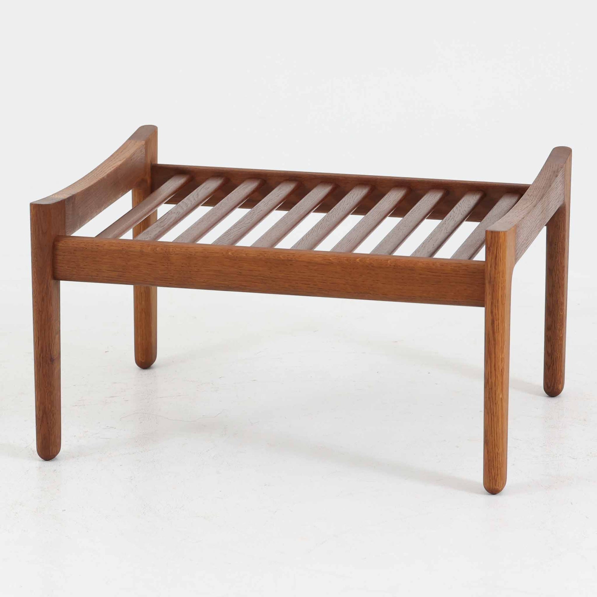 ハンス・J・ウェグナーがデザインし、ゲタマ社から発表されたGE290オットマンは、堅牢なオーク材のフレームと、ウール混紡のざっくりとした風合いを持つダークグレーの張地が美しく調和した北欧ビンテージ家具です。直線的でありながら丸みを帯びた脚部が軽快な印象を与え、ソファと組み合わせてフットレストとして、あるいは単体で腰掛けとしてもお使いいただけます。オーク材の色味は少し濃いめで、本革カバー仕様のビンテージモデルのためクッション内部にスプリングは元々内包されないタイプです。経年によって深まった木肌の風合いと上質な張地が、空間に落ち着いた存在感をもたらす逸品です。