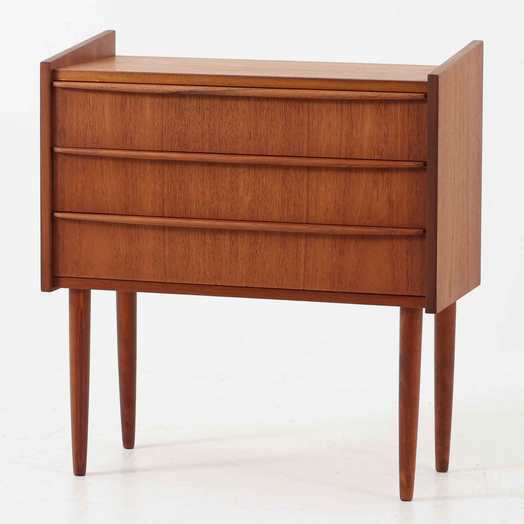 チーク材を使用したデンマーク製の3段チェストで、直線的な本体に対して丸みを帯びた引き手と細身のテーパードレッグが柔らかな印象を与え、さらに天板左右に伸びた耳のような張り出しがデザインのアクセントとなり、北欧らしい優雅な佇まいが感じられます。引き出しは3杯備え、衣類や小物収納に適したサイズ感で、ベッドサイドやリビングの一角に置きやすく、花器や照明のディスプレイ台としても楽しめます。シンプルな造形ながら木目の美しさが際立ち、空間に温かみと落ち着きを添えてくれるビンテージ家具です。