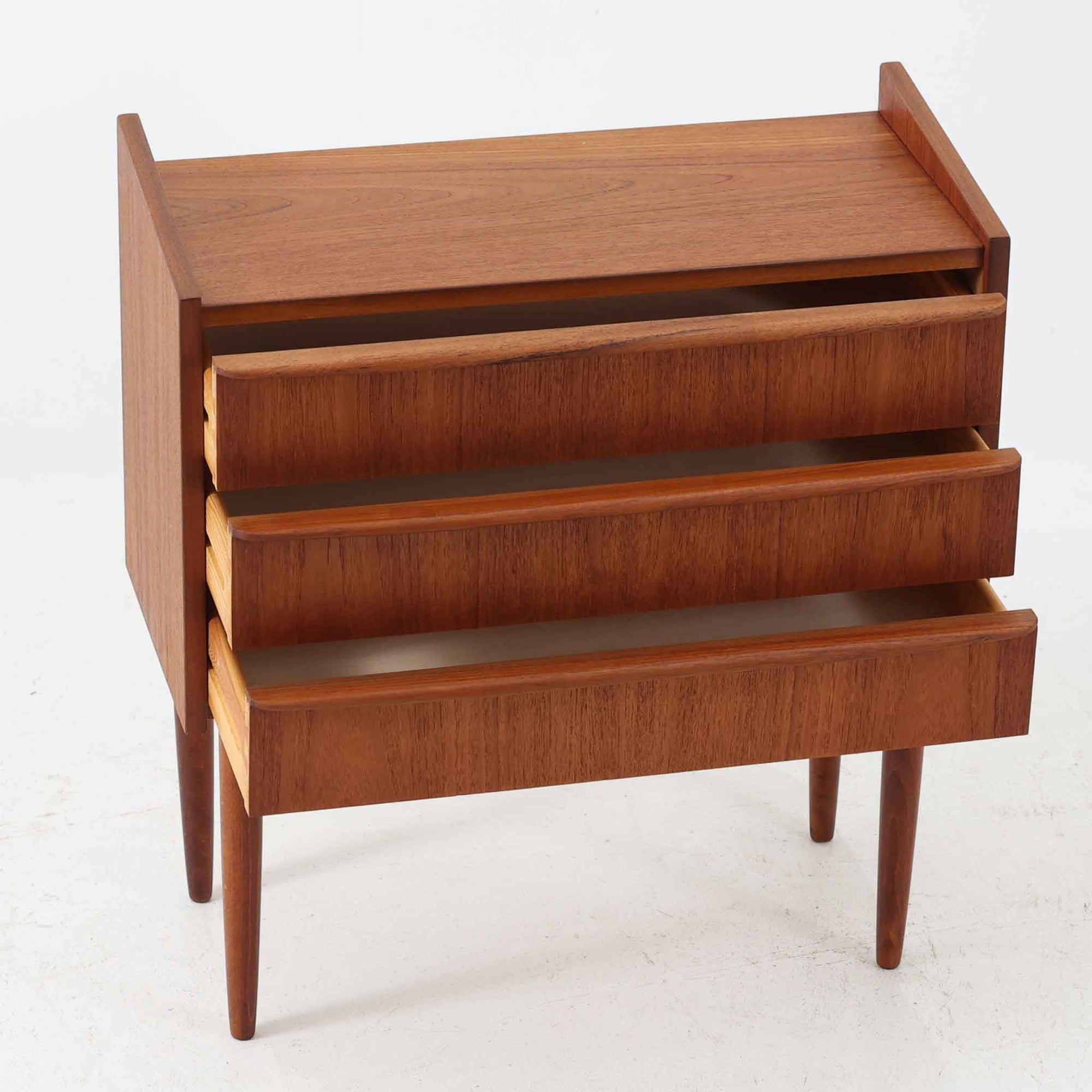 チーク材を使用したデンマーク製の3段チェストで、直線的な本体に対して丸みを帯びた引き手と細身のテーパードレッグが柔らかな印象を与え、さらに天板左右に伸びた耳のような張り出しがデザインのアクセントとなり、北欧らしい優雅な佇まいが感じられます。引き出しは3杯備え、衣類や小物収納に適したサイズ感で、ベッドサイドやリビングの一角に置きやすく、花器や照明のディスプレイ台としても楽しめます。シンプルな造形ながら木目の美しさが際立ち、空間に温かみと落ち着きを添えてくれるビンテージ家具です。