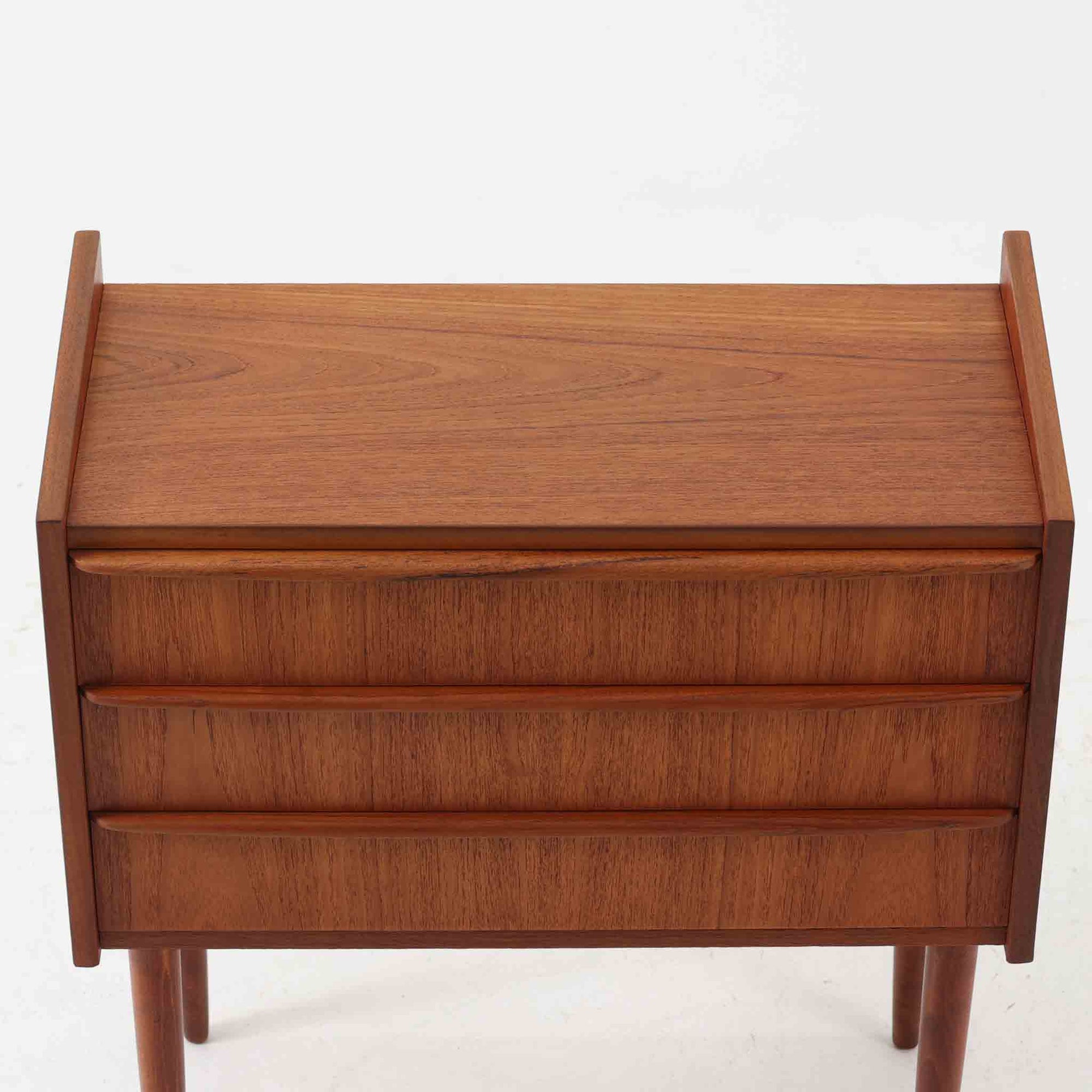 チーク材を使用したデンマーク製の3段チェストで、直線的な本体に対して丸みを帯びた引き手と細身のテーパードレッグが柔らかな印象を与え、さらに天板左右に伸びた耳のような張り出しがデザインのアクセントとなり、北欧らしい優雅な佇まいが感じられます。引き出しは3杯備え、衣類や小物収納に適したサイズ感で、ベッドサイドやリビングの一角に置きやすく、花器や照明のディスプレイ台としても楽しめます。シンプルな造形ながら木目の美しさが際立ち、空間に温かみと落ち着きを添えてくれるビンテージ家具です。