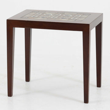 Severin Hansen Jr.（セヴェリン・ハンセン）がデザインし、Haslev社より製造されたタイルトップテーブルで、天板にはNils ThorssonによるBACAシリーズの陶板が贅沢にあしらわれています。端正な角脚を持つマホガニー材のフレームは、ハンセンらしいシャープなジョイントが際立ち、凛とした佇まいを演出します。耐熱性に優れたタイルは日常使いに適しており、サイドテーブルや植物のディスプレイスペースとしても活躍。経年により深みを増した木肌とBACA特有の芸術性が空間に上質なアクセントを添えるビンテージ品です。