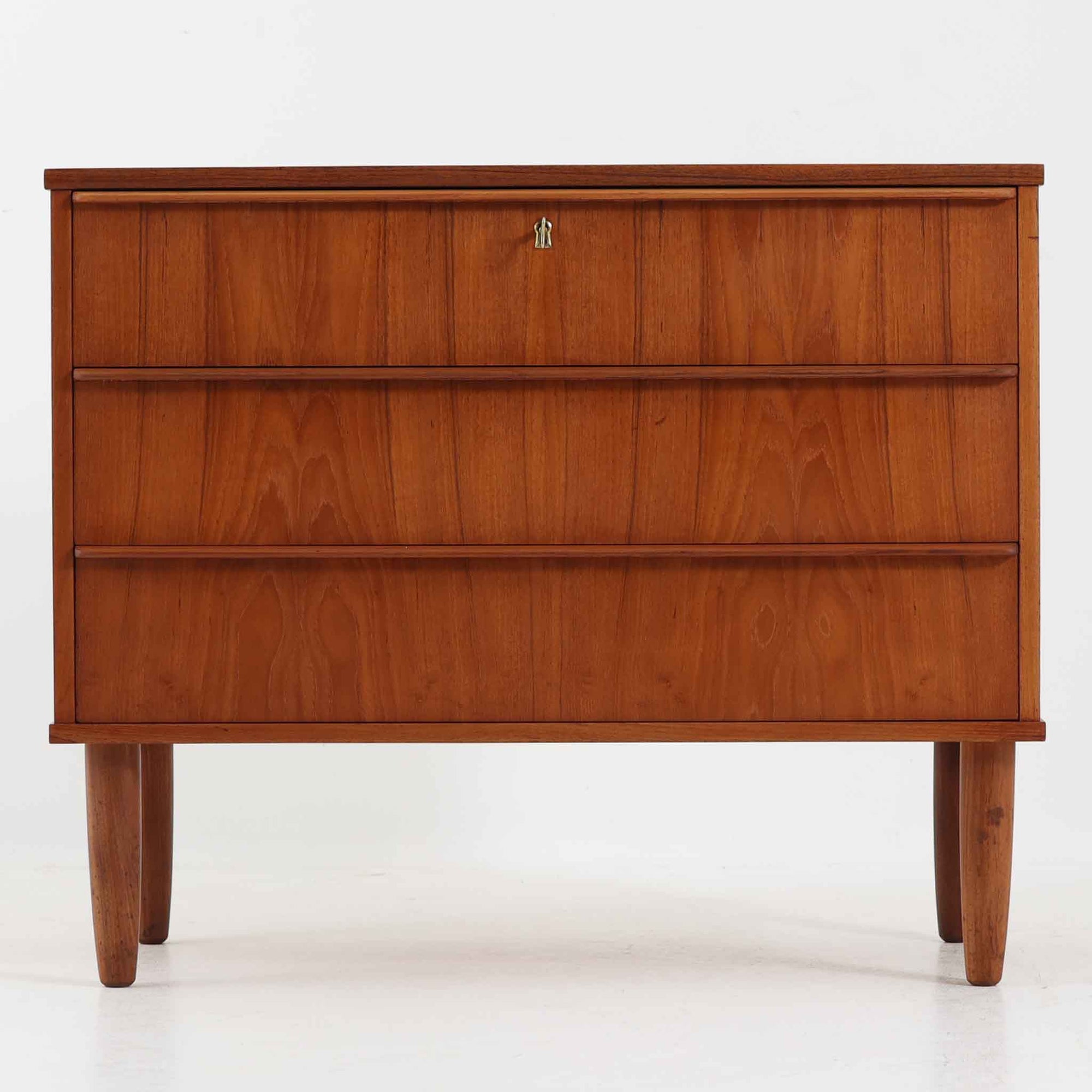 デンマーク製のチーク材3段チェストです。前板の一体的なデザインが美しく、取手部分を削り出した滑らかなラインが上品な印象を与えます。幅が77cmと少し大きめで、コンパクトながら収納力があり、寝室や玄関、リビングのサイド収納としても活躍します。最上段には鍵付きの引き出しが設けられ、大切な書類や貴重品の保管にも便利です。脚部まで丁寧に仕上げられた造りは北欧家具ならではの職人技を感じさせ、空間に温もりを添えてくれます。