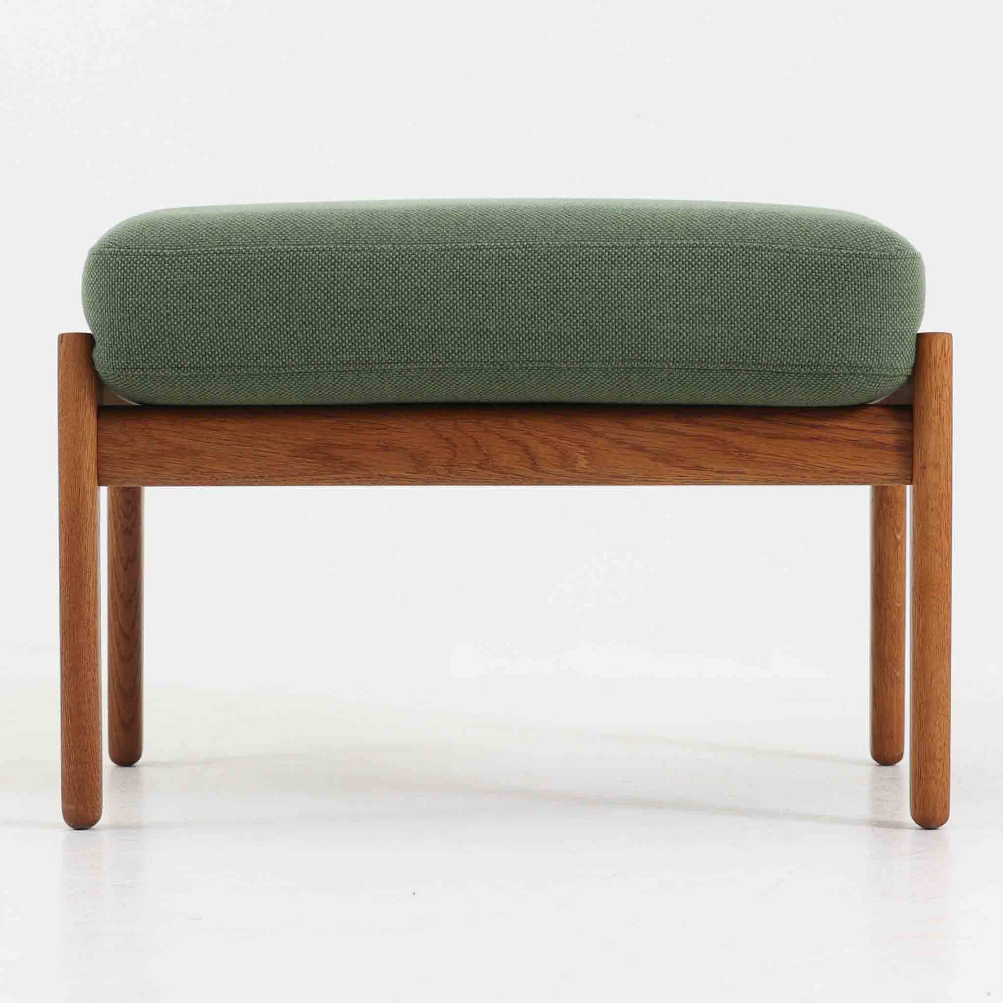 ハンス・J・ウェグナーがデザインし、ゲタマ社から発表されたGE290オットマンは、堅牢なオーク材のフレームと、ウール混紡のざっくりとした風合いを持つ落ち着いたグリーン系の張地が美しく調和した北欧ビンテージ家具です。直線的でありながら丸みを帯びた脚部が軽快な印象を与え、ソファと組み合わせてフットレストとして、あるいは単体で腰掛けとしてもお使いいただけます。経年によって深まった木肌の風合いと上質な張地が空間に落ち着いた存在感をもたらし、クッション内部にはビンテージ品ならではのスプリングが内包されています。