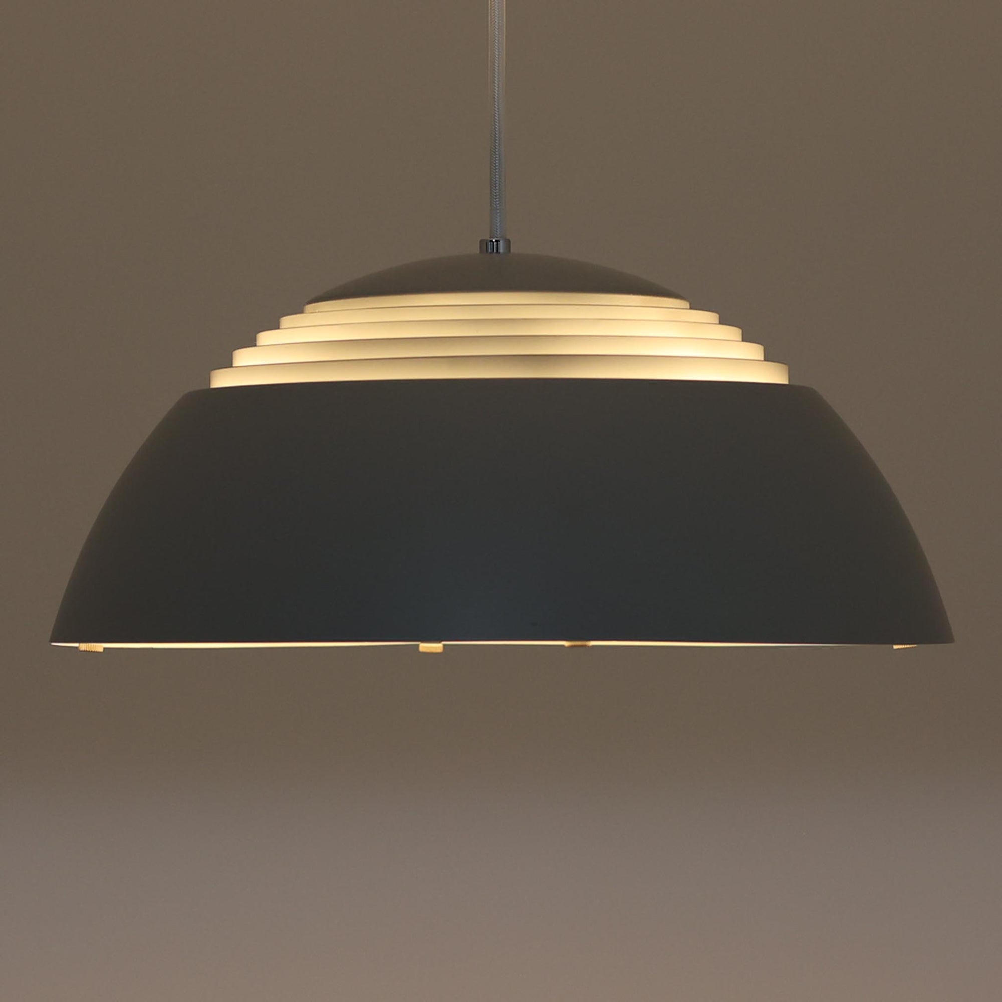 Arne JacobsenがLouis Poulsenのためにデザインした「AJ Royal 370」ペンダントライトです。直径37cmのサイズで、段階的に重なる上部シェードと、下面を覆うアクリルディフューザーによって光が柔らかく拡散し、均一で心地よい明かりを提供します。アルミとスチールの堅牢な構造に加え、シンプルかつモダンなフォルムが空間に洗練された印象を与えます。ダイニングやリビングに適した存在感ある照明で、北欧デザインを象徴するビンテージアイテムです。現行モデルでは展開されていないライトグレーのカラーは、当時のオリジナル品ならではの希少な仕様であり、コレクターズアイテムとしても高い価値を持っています。