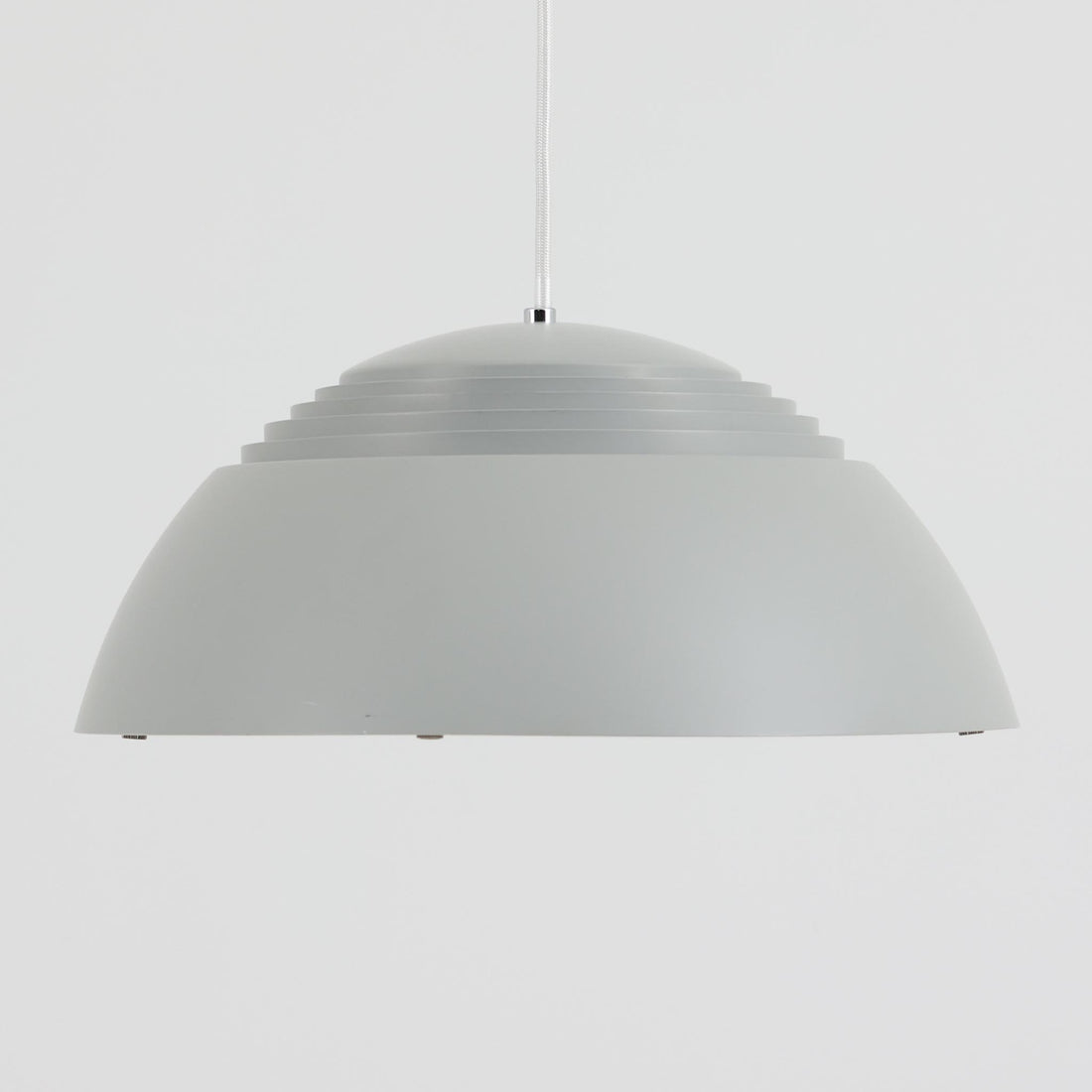 Arne JacobsenがLouis Poulsenのためにデザインした「AJ Royal 370」ペンダントライトです。直径37cmのサイズで、段階的に重なる上部シェードと、下面を覆うアクリルディフューザーによって光が柔らかく拡散し、均一で心地よい明かりを提供します。アルミとスチールの堅牢な構造に加え、シンプルかつモダンなフォルムが空間に洗練された印象を与えます。ダイニングやリビングに適した存在感ある照明で、北欧デザインを象徴するビンテージアイテムです。現行モデルでは展開されていないライトグレーのカラーは、当時のオリジナル品ならではの希少な仕様であり、コレクターズアイテムとしても高い価値を持っています。