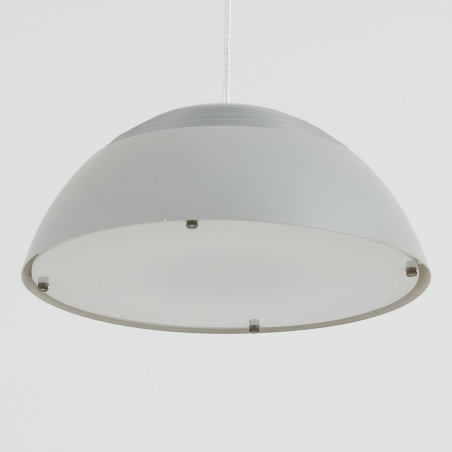 Arne JacobsenがLouis Poulsenのためにデザインした「AJ Royal 370」ペンダントライトです。直径37cmのサイズで、段階的に重なる上部シェードと、下面を覆うアクリルディフューザーによって光が柔らかく拡散し、均一で心地よい明かりを提供します。アルミとスチールの堅牢な構造に加え、シンプルかつモダンなフォルムが空間に洗練された印象を与えます。ダイニングやリビングに適した存在感ある照明で、北欧デザインを象徴するビンテージアイテムです。現行モデルでは展開されていないライトグレーのカラーは、当時のオリジナル品ならではの希少な仕様であり、コレクターズアイテムとしても高い価値を持っています。