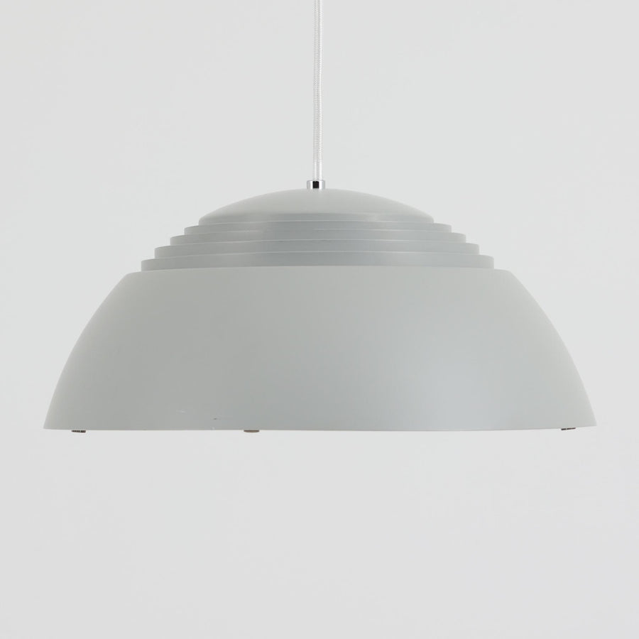Arne JacobsenがLouis Poulsenのためにデザインした「AJ Royal 370」ペンダントライトです。直径37cmのサイズで、段階的に重なる上部シェードと、下面を覆うアクリルディフューザーによって光が柔らかく拡散し、均一で心地よい明かりを提供します。アルミとスチールの堅牢な構造に加え、シンプルかつモダンなフォルムが空間に洗練された印象を与えます。ダイニングやリビングに適した存在感ある照明で、北欧デザインを象徴するビンテージアイテムです。現行モデルでは展開されていないライトグレーのカラーは、当時のオリジナル品ならではの希少な仕様であり、コレクターズアイテムとしても高い価値を持っています。