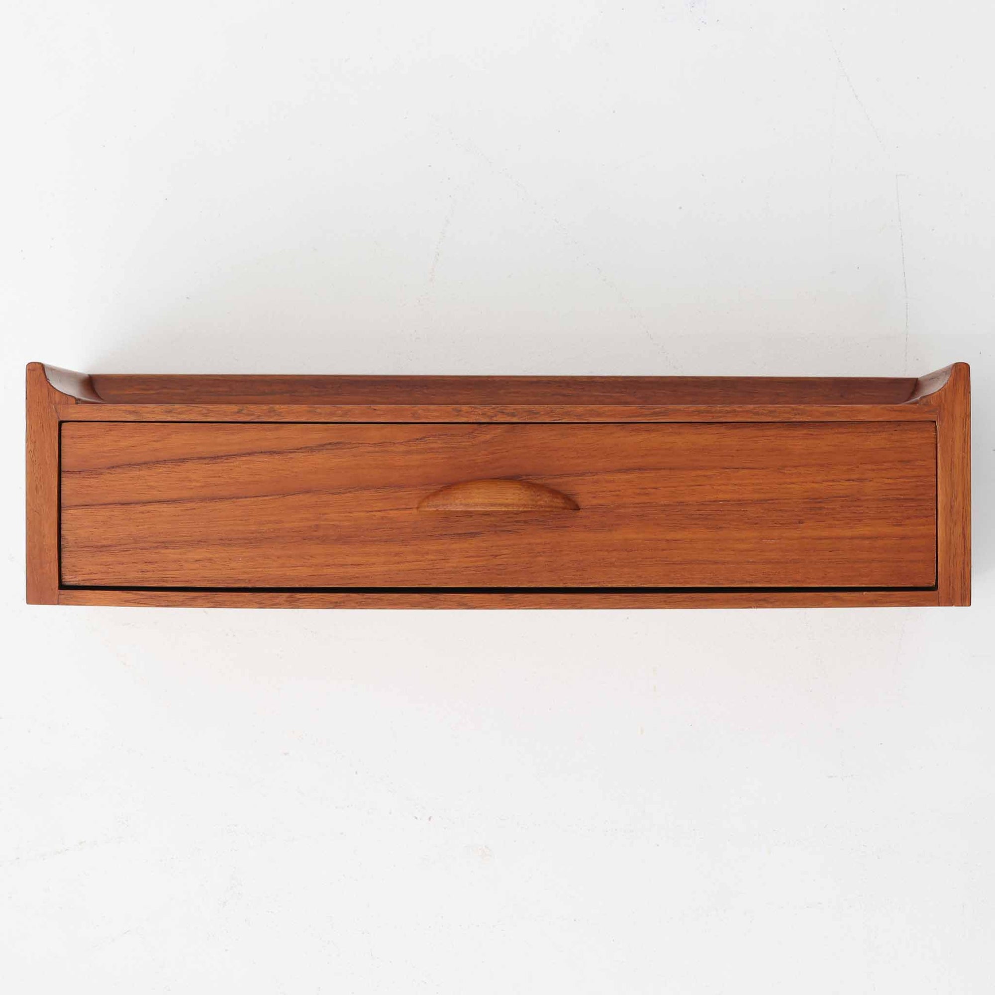 チーク材を使用したデンマーク製のウォールシェルフは、壁付けタイプならではの省スペース性に加え、滑らかな木目と美しい色合いが空間に温もりを添える北欧ビンテージ家具です。引き出し付きの設計により、鍵や文具、小物の収納に最適で、玄関やベッドサイドにも馴染みます。天板には立ち上がりが設けられており、ディスプレイスペースとしても活用可能です。時を重ねた木肌の味わいが一点物ならではの魅力を醸し出しています。