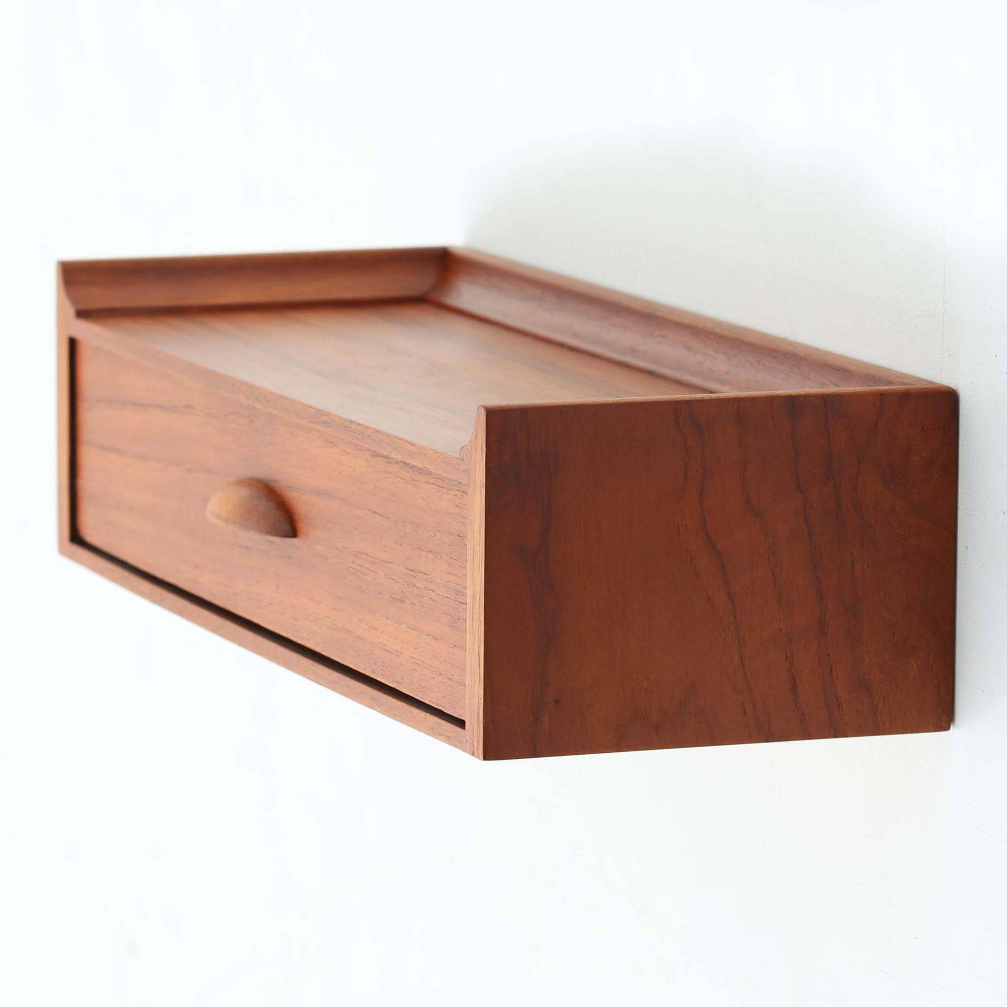 チーク材を使用したデンマーク製のウォールシェルフは、壁付けタイプならではの省スペース性に加え、滑らかな木目と美しい色合いが空間に温もりを添える北欧ビンテージ家具です。引き出し付きの設計により、鍵や文具、小物の収納に最適で、玄関やベッドサイドにも馴染みます。天板には立ち上がりが設けられており、ディスプレイスペースとしても活用可能です。時を重ねた木肌の味わいが一点物ならではの魅力を醸し出しています。