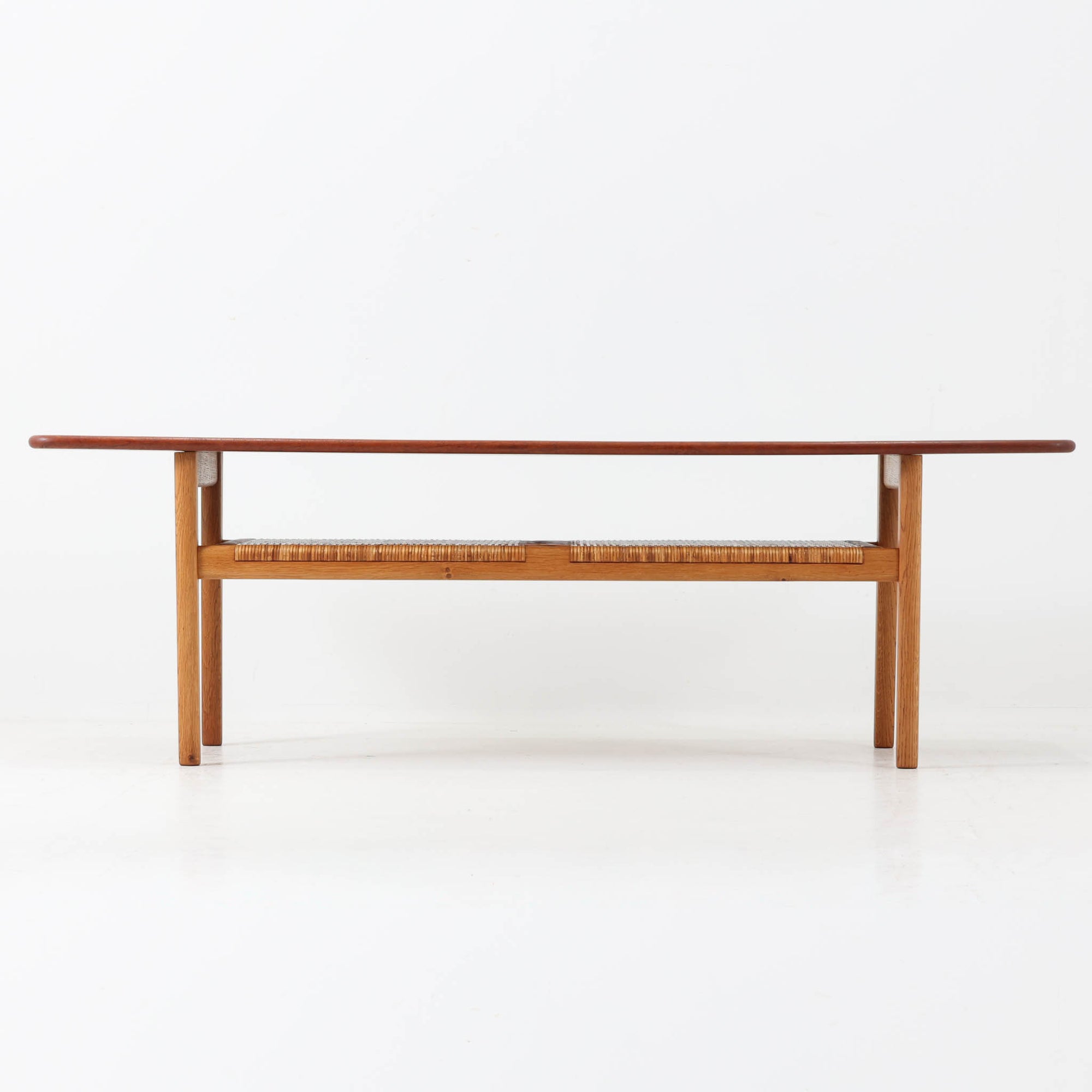 Hans J. WegnerがAndreas Tuckのために手掛けたAT10センターテーブルは、チーク材の天板にオーク材のフレーム、ラタン張りの棚板を組み合わせた、素材使いの美しさが際立つ北欧ビンテージの名作です。直線を基調とした端正なプロポーションの中に、天板縁のやわらかな丸みや、軽やかに抜け感を生むラタン棚が配され、視覚的にも圧迫感のない佇まいに仕上げられています。幅160cmのゆとりあるサイズはリビングの主役として使いやすく、雑誌や小物を収納できる実用性も備え、経年変化により深みを増した木肌が空間に上質な落ち着きをもたらします。なお、天板には反りがあり、大きめのへこみも見られるため、その点を考慮した価格設定としておりますが、ご使用には問題なく、ビンテージならではの風合いとしてご理解いただける方におすすめの一台です。