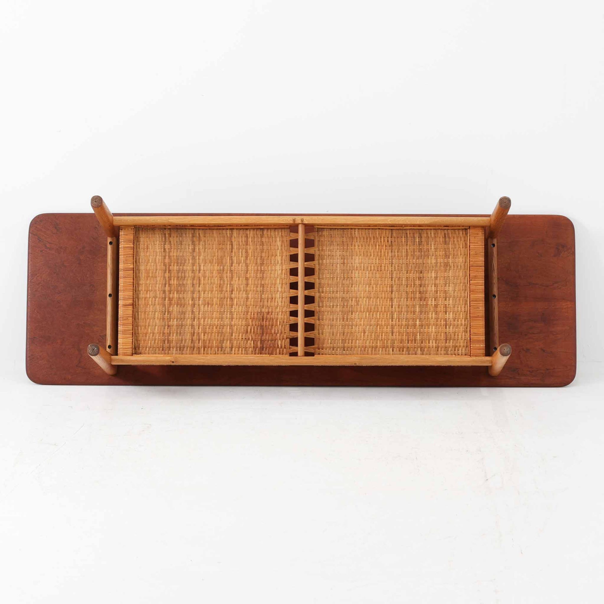 Hans J. WegnerがAndreas Tuckのために手掛けたAT10センターテーブルは、チーク材の天板にオーク材のフレーム、ラタン張りの棚板を組み合わせた、素材使いの美しさが際立つ北欧ビンテージの名作です。直線を基調とした端正なプロポーションの中に、天板縁のやわらかな丸みや、軽やかに抜け感を生むラタン棚が配され、視覚的にも圧迫感のない佇まいに仕上げられています。幅160cmのゆとりあるサイズはリビングの主役として使いやすく、雑誌や小物を収納できる実用性も備え、経年変化により深みを増した木肌が空間に上質な落ち着きをもたらします。なお、天板には反りがあり、大きめのへこみも見られるため、その点を考慮した価格設定としておりますが、ご使用には問題なく、ビンテージならではの風合いとしてご理解いただける方におすすめの一台です。