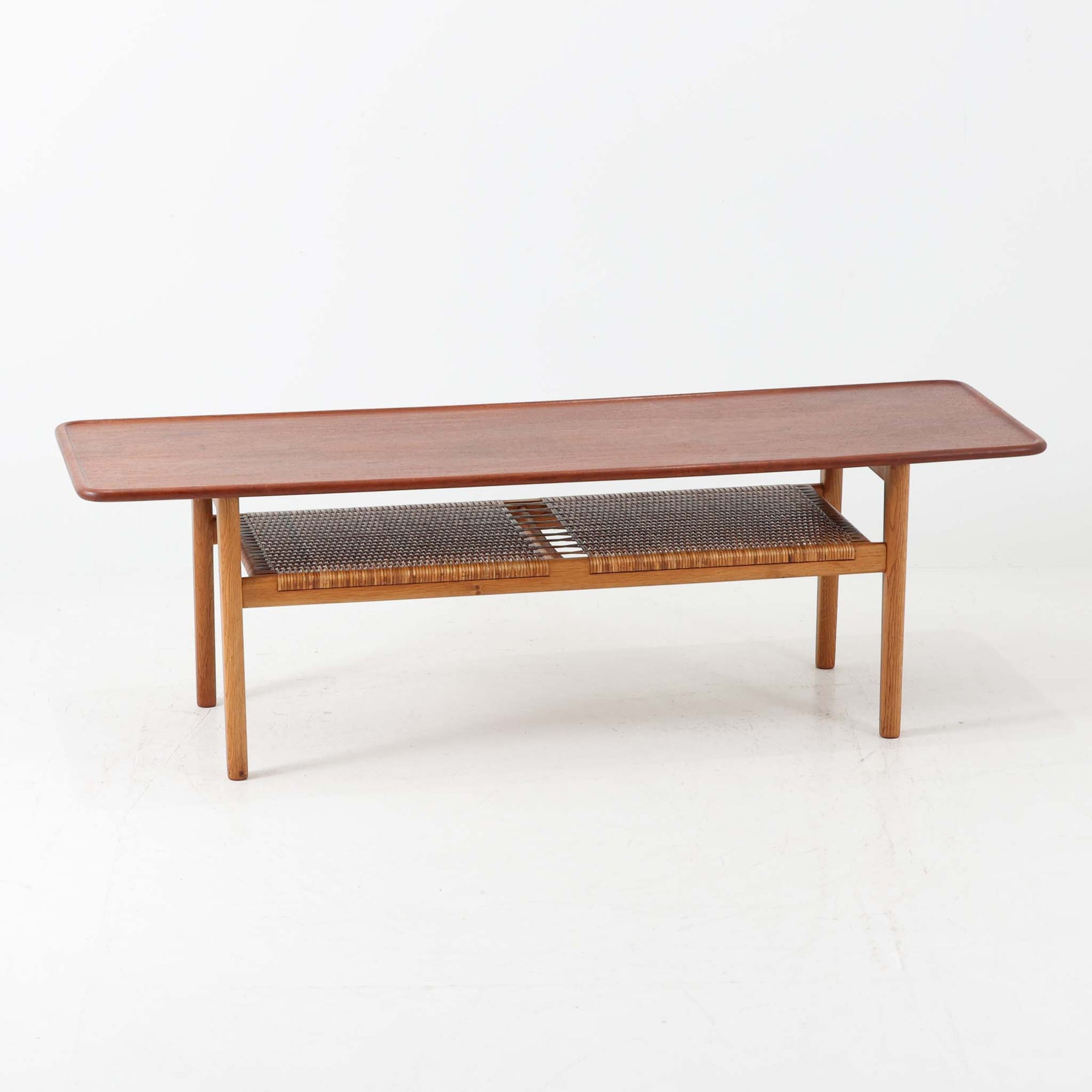 Hans J. WegnerがAndreas Tuckのために手掛けたAT10センターテーブルは、チーク材の天板にオーク材のフレーム、ラタン張りの棚板を組み合わせた、素材使いの美しさが際立つ北欧ビンテージの名作です。直線を基調とした端正なプロポーションの中に、天板縁のやわらかな丸みや、軽やかに抜け感を生むラタン棚が配され、視覚的にも圧迫感のない佇まいに仕上げられています。幅160cmのゆとりあるサイズはリビングの主役として使いやすく、雑誌や小物を収納できる実用性も備え、経年変化により深みを増した木肌が空間に上質な落ち着きをもたらします。なお、天板には反りがあり、大きめのへこみも見られるため、その点を考慮した価格設定としておりますが、ご使用には問題なく、ビンテージならではの風合いとしてご理解いただける方におすすめの一台です。