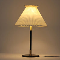 Aage Petersenによるデザインで、Le Klint社より発表されたモデル352のテーブルランプです。高さ62～85cmの昇降機能を備え、使用シーンに合わせて調整可能なのが魅力です。黒塗装のスチール支柱に真鍮パーツがアクセントとなり、プリーツ加工が美しい紙製シェードからは柔らかな光が広がります。特に点灯時にはシェードを透過して漏れる灯りが印象的で、空間を上品に演出します。現在は廃盤となっており、ビンテージ品のみ入手可能なモデルです。