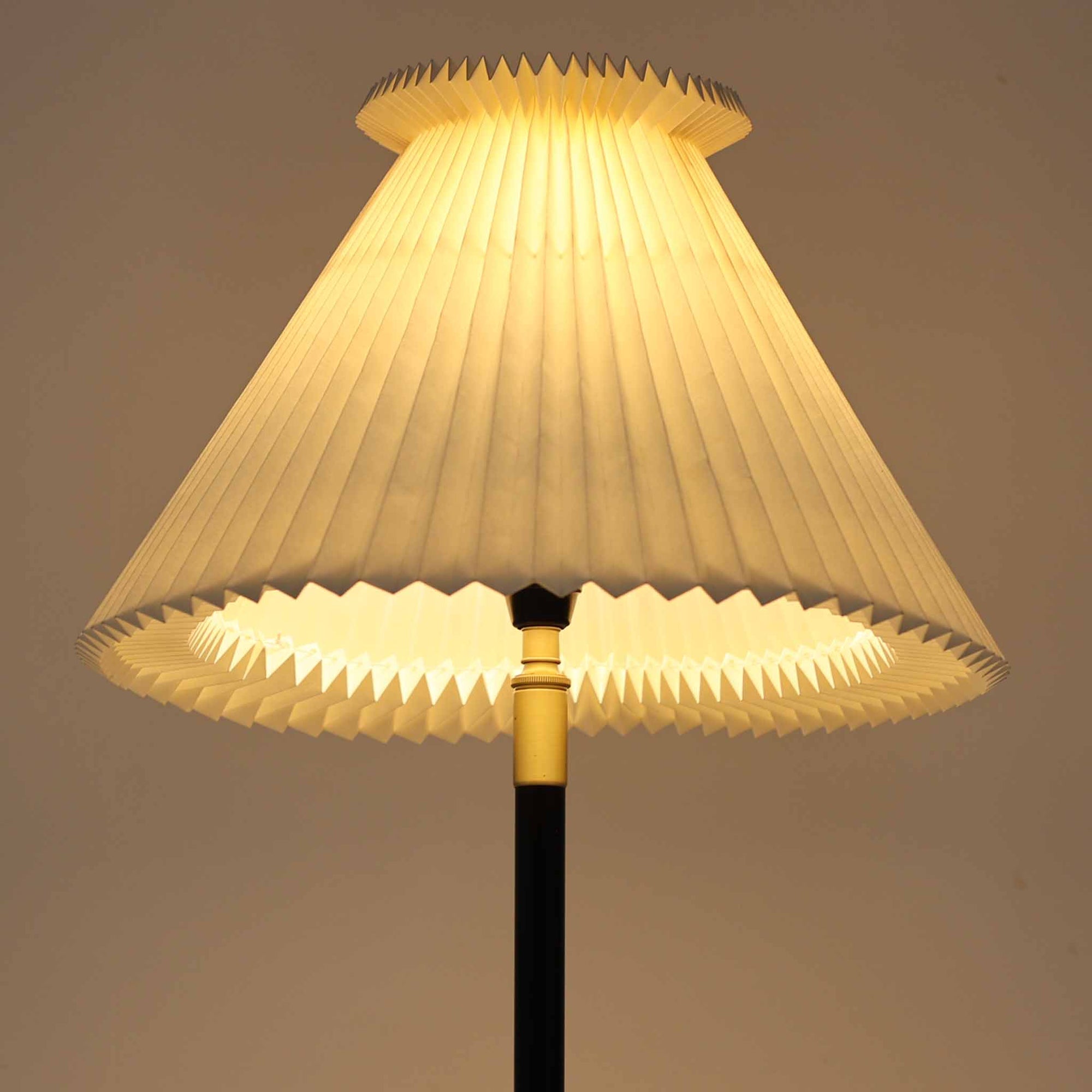 Aage Petersenによるデザインで、Le Klint社より発表されたモデル352のテーブルランプです。高さ62～85cmの昇降機能を備え、使用シーンに合わせて調整可能なのが魅力です。黒塗装のスチール支柱に真鍮パーツがアクセントとなり、プリーツ加工が美しい紙製シェードからは柔らかな光が広がります。特に点灯時にはシェードを透過して漏れる灯りが印象的で、空間を上品に演出します。現在は廃盤となっており、ビンテージ品のみ入手可能なモデルです。