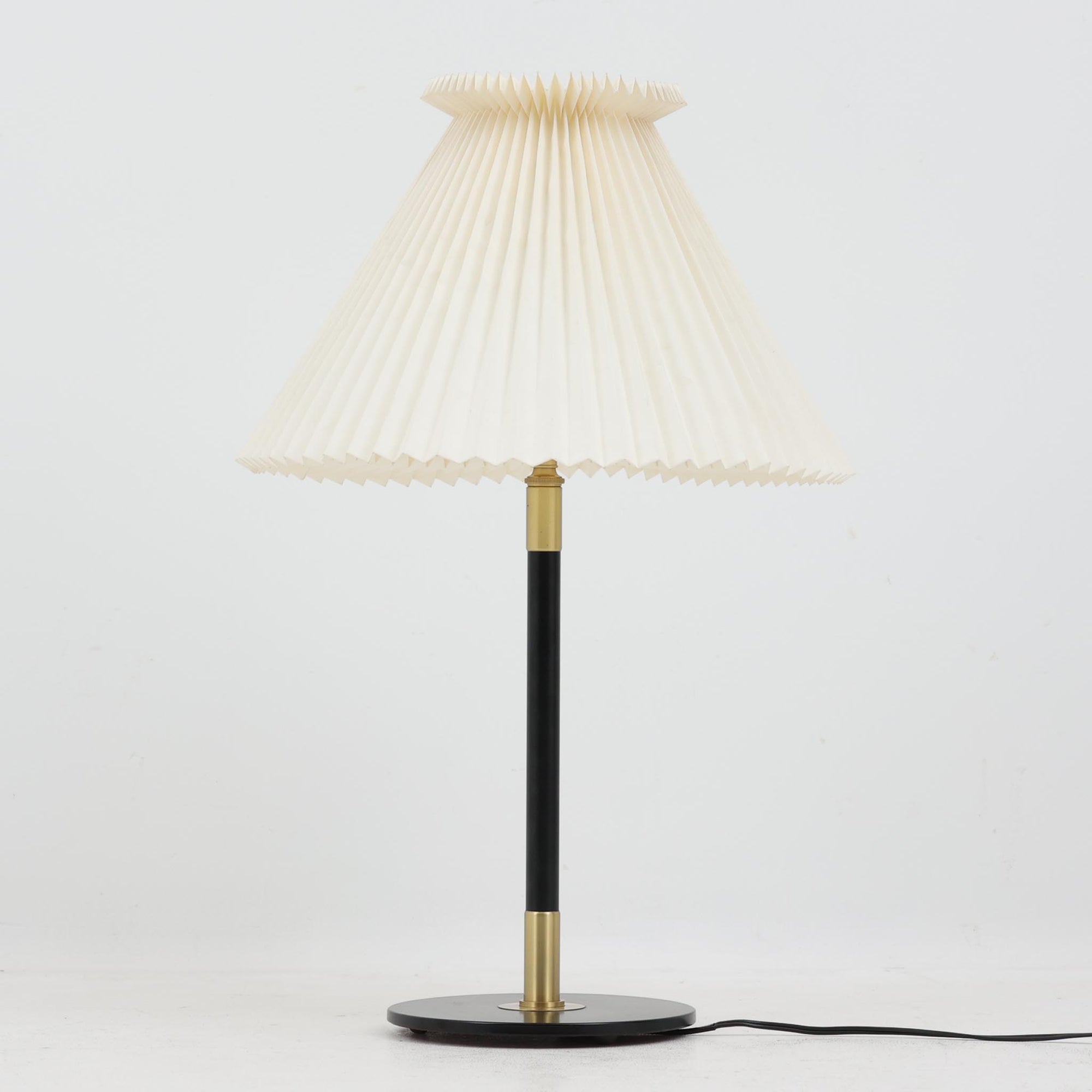 Aage Petersenによるデザインで、Le Klint社より発表されたモデル352のテーブルランプです。高さ62～85cmの昇降機能を備え、使用シーンに合わせて調整可能なのが魅力です。黒塗装のスチール支柱に真鍮パーツがアクセントとなり、プリーツ加工が美しい紙製シェードからは柔らかな光が広がります。特に点灯時にはシェードを透過して漏れる灯りが印象的で、空間を上品に演出します。現在は廃盤となっており、ビンテージ品のみ入手可能なモデルです。