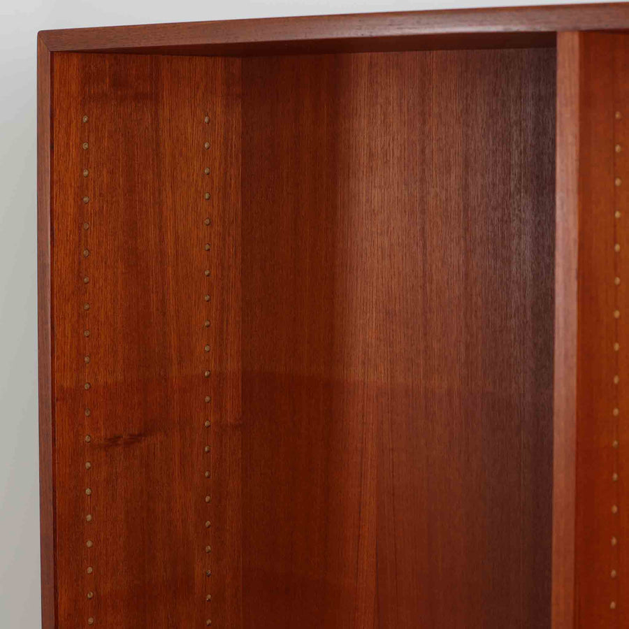 デンマークを代表するデザイナー、Borge Mogensen（ボーエ・モーエンセン）によるFDB社製のチーク材ブックシェルフです。無駄を削ぎ落とした直線的なフォルムと、シンプルで機能的なデザインがモーエンセンらしい魅力を感じさせます。棚板は高さ調整が可能で、収納するものに合わせて柔軟にレイアウトを変えることができます。チークの豊かな木目と深みのある色合いが上品な印象を与え、経年により増した艶や質感も味わい深い一点です。台輪部分までチーク材が使用された希少なタイプで、素材の統一感と高級感が際立ちます。リビングでの書籍収納はもちろん、オブジェや陶器を飾るディスプレイシェルフとしても活躍する北欧ビンテージ家具です。