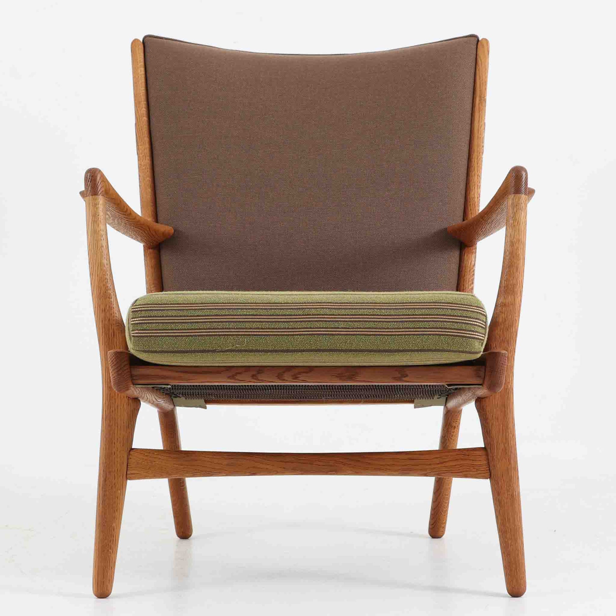 Hans J. Wegner（ハンス・J・ウェグナー）によるイージーチェア「AP16」。デンマークの名門AP stolen社より製造された一脚です。緩やかに傾斜した背もたれと、滑らかに削り出されたアームの曲線が美しく調和し、オーク材の力強い木目と職人技が際立ちます。背面にはブラウン、座面にはグリーンストライプの張地を採用し、上品ながらも温かみのある印象を与えます。現地デンマークのオークションで入手した確かな出自を持つ本品は、読書やくつろぎの時間をより豊かなものにしてくれます。経年による木部の風合いがさらに深みを加えています。