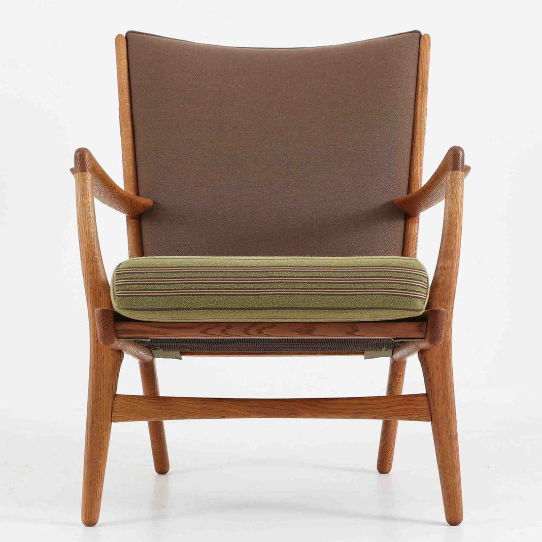 Hans J. Wegner（ハンス・J・ウェグナー）によるイージーチェア「AP16」。デンマークの名門AP stolen社より製造された一脚です。緩やかに傾斜した背もたれと、滑らかに削り出されたアームの曲線が美しく調和し、オーク材の力強い木目と職人技が際立ちます。背面にはブラウン、座面にはグリーンストライプの張地を採用し、上品ながらも温かみのある印象を与えます。現地デンマークのオークションで入手した確かな出自を持つ本品は、読書やくつろぎの時間をより豊かなものにしてくれます。経年による木部の風合いがさらに深みを加えています。