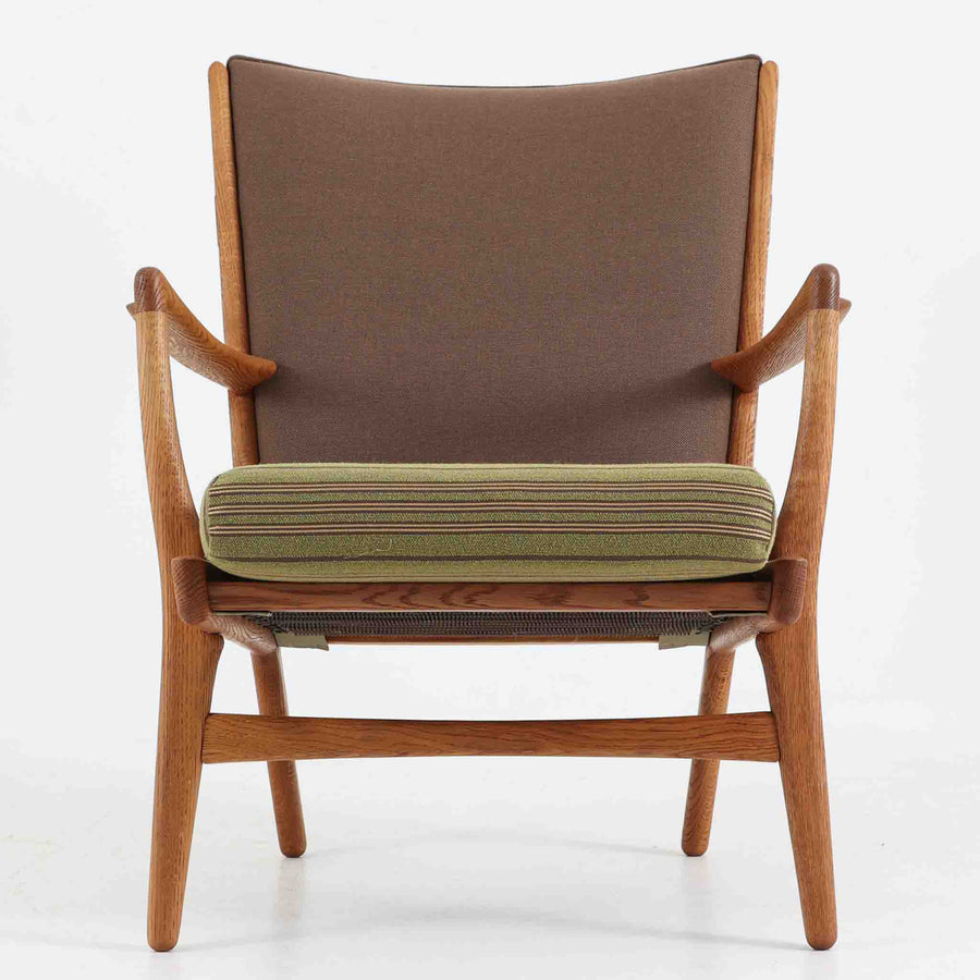 Hans J. Wegner（ハンス・J・ウェグナー）によるイージーチェア「AP16」。デンマークの名門AP stolen社より製造された一脚です。緩やかに傾斜した背もたれと、滑らかに削り出されたアームの曲線が美しく調和し、オーク材の力強い木目と職人技が際立ちます。背面にはブラウン、座面にはグリーンストライプの張地を採用し、上品ながらも温かみのある印象を与えます。現地デンマークのオークションで入手した確かな出自を持つ本品は、読書やくつろぎの時間をより豊かなものにしてくれます。経年による木部の風合いがさらに深みを加えています。
