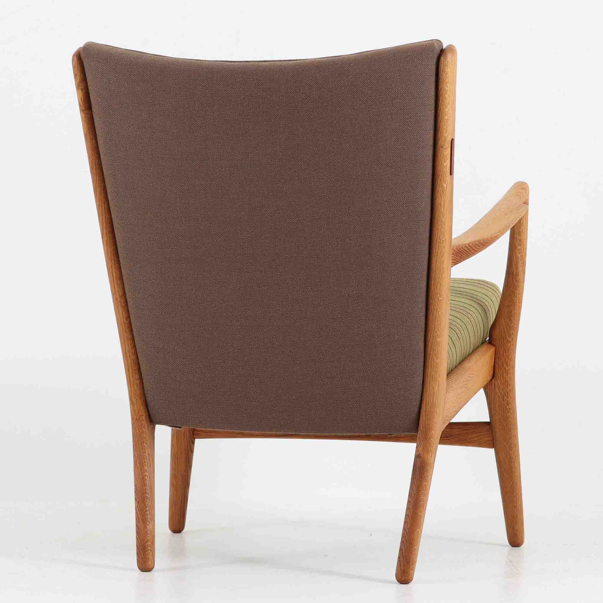 Hans J. Wegner（ハンス・J・ウェグナー）によるイージーチェア「AP16」。デンマークの名門AP stolen社より製造された一脚です。緩やかに傾斜した背もたれと、滑らかに削り出されたアームの曲線が美しく調和し、オーク材の力強い木目と職人技が際立ちます。背面にはブラウン、座面にはグリーンストライプの張地を採用し、上品ながらも温かみのある印象を与えます。現地デンマークのオークションで入手した確かな出自を持つ本品は、読書やくつろぎの時間をより豊かなものにしてくれます。経年による木部の風合いがさらに深みを加えています。