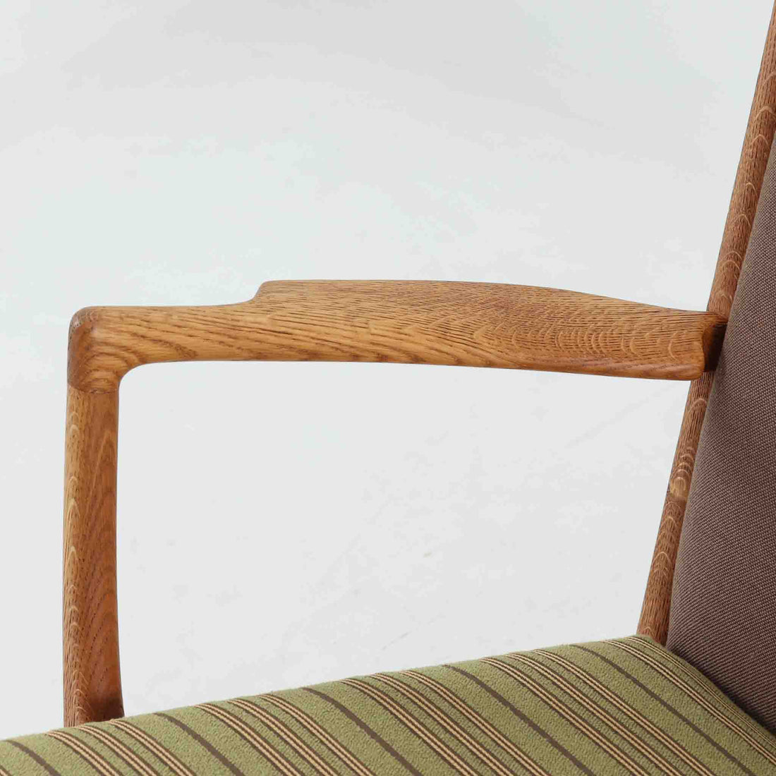 Hans J. Wegner（ハンス・J・ウェグナー）によるイージーチェア「AP16」。デンマークの名門AP stolen社より製造された一脚です。緩やかに傾斜した背もたれと、滑らかに削り出されたアームの曲線が美しく調和し、オーク材の力強い木目と職人技が際立ちます。背面にはブラウン、座面にはグリーンストライプの張地を採用し、上品ながらも温かみのある印象を与えます。現地デンマークのオークションで入手した確かな出自を持つ本品は、読書やくつろぎの時間をより豊かなものにしてくれます。経年による木部の風合いがさらに深みを加えています。