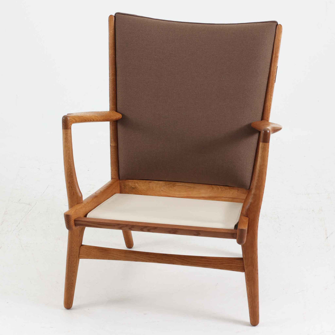 Hans J. Wegner（ハンス・J・ウェグナー）によるイージーチェア「AP16」。デンマークの名門AP stolen社より製造された一脚です。緩やかに傾斜した背もたれと、滑らかに削り出されたアームの曲線が美しく調和し、オーク材の力強い木目と職人技が際立ちます。背面にはブラウン、座面にはグリーンストライプの張地を採用し、上品ながらも温かみのある印象を与えます。現地デンマークのオークションで入手した確かな出自を持つ本品は、読書やくつろぎの時間をより豊かなものにしてくれます。経年による木部の風合いがさらに深みを加えています。